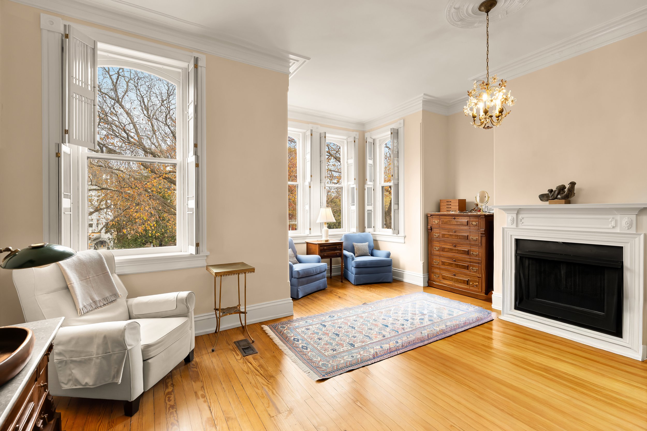 Bright living room with hardwood floors, large windows with shutters, a white armchair with a blanket, a rug, a wooden chest of drawers, two blue armchairs, a lamp, and a chandelier.