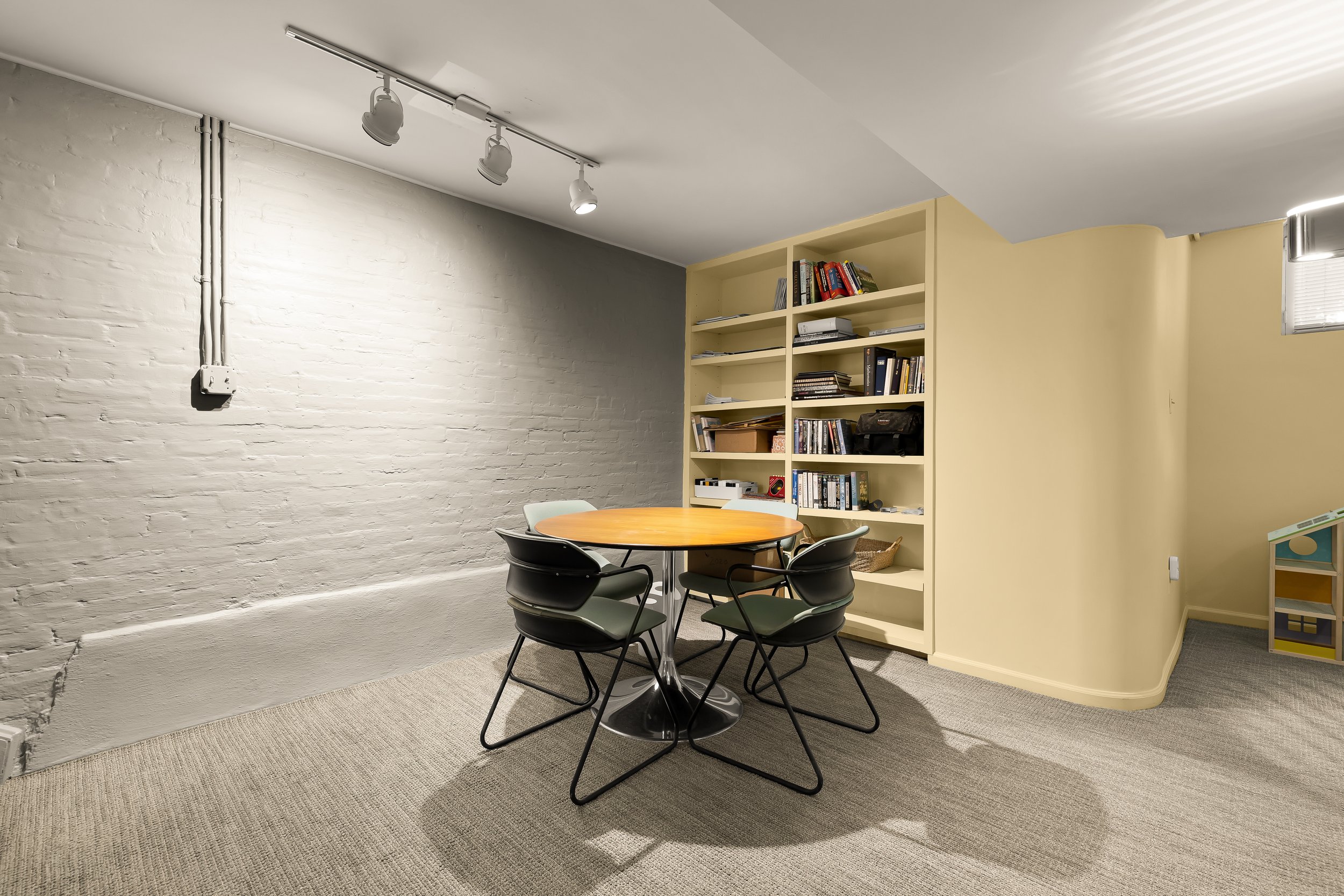 Undersized basement room with a small round table, five chairs, a cream bookshelf filled with books and items, a white brick wall, and a window with blinds.