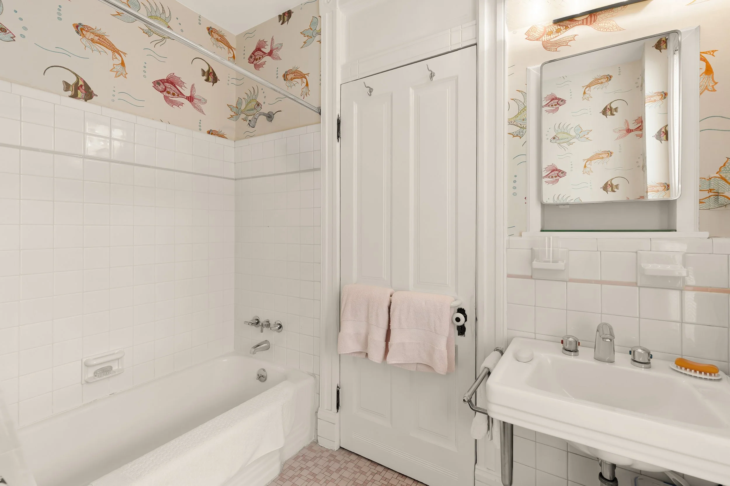 A bathroom with white tiled walls and a bathtubs, featuring a wallpaper border with colorful fish on the upper wall. There is a mirror above the sink, and a small white sink with a faucet and two knobs. A pink towel hangs on a rack on a door next to 