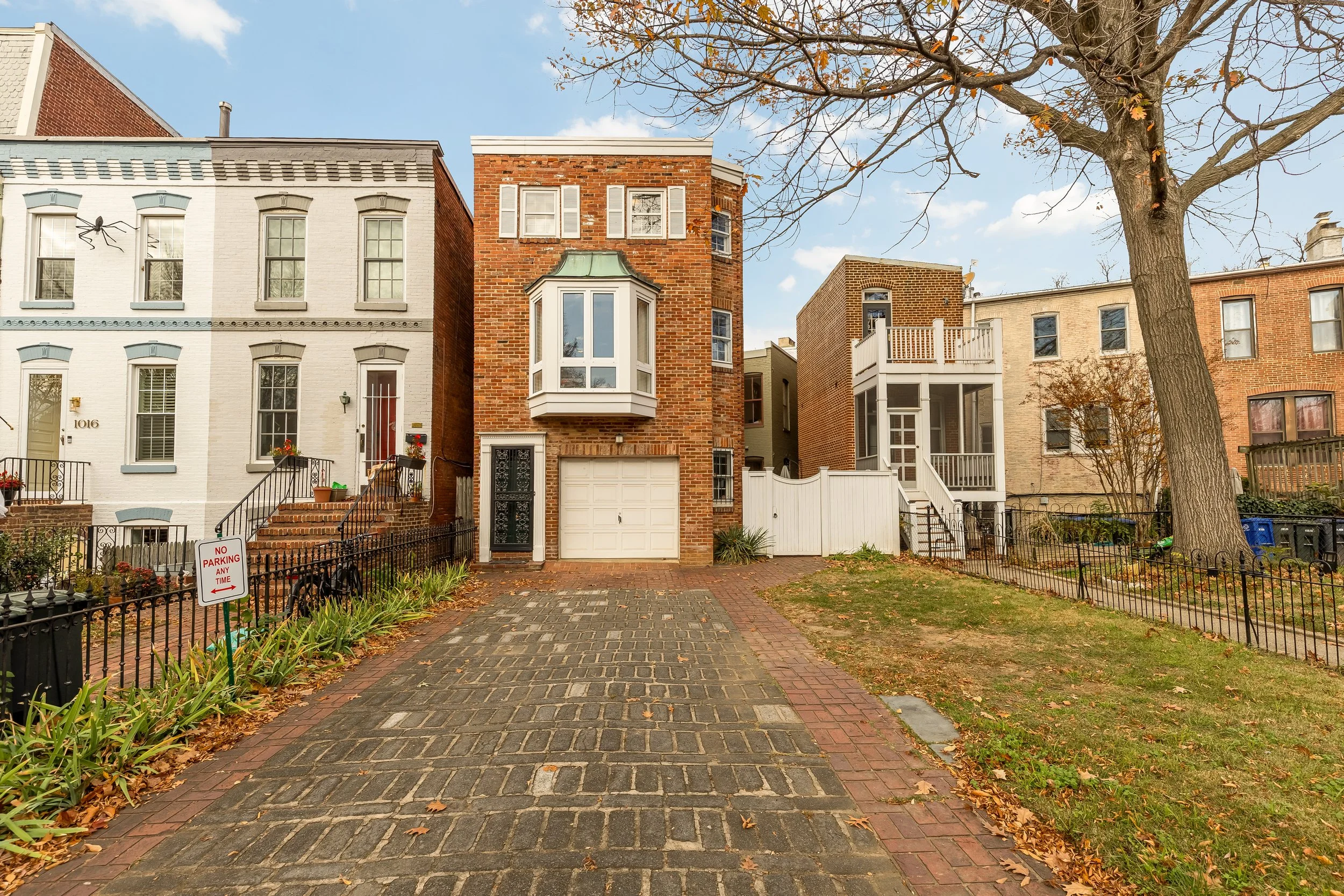 Residential street view with brick and white houses, a driveway, a large tree, and a no parking sign, taken during fall.