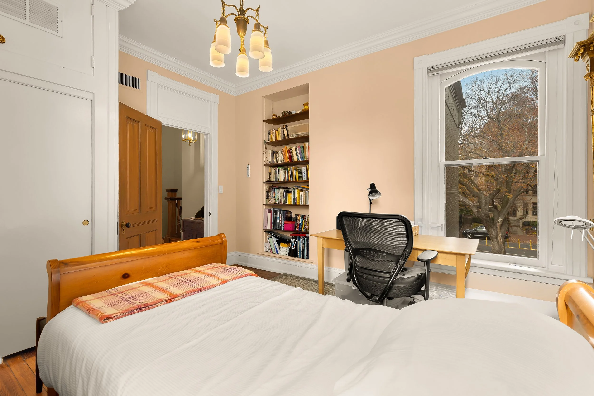 A bedroom with a wooden bed, a desk with a black office chair, a built-in bookshelf filled with books, a large window showing trees outside, and a ceiling light fixture.