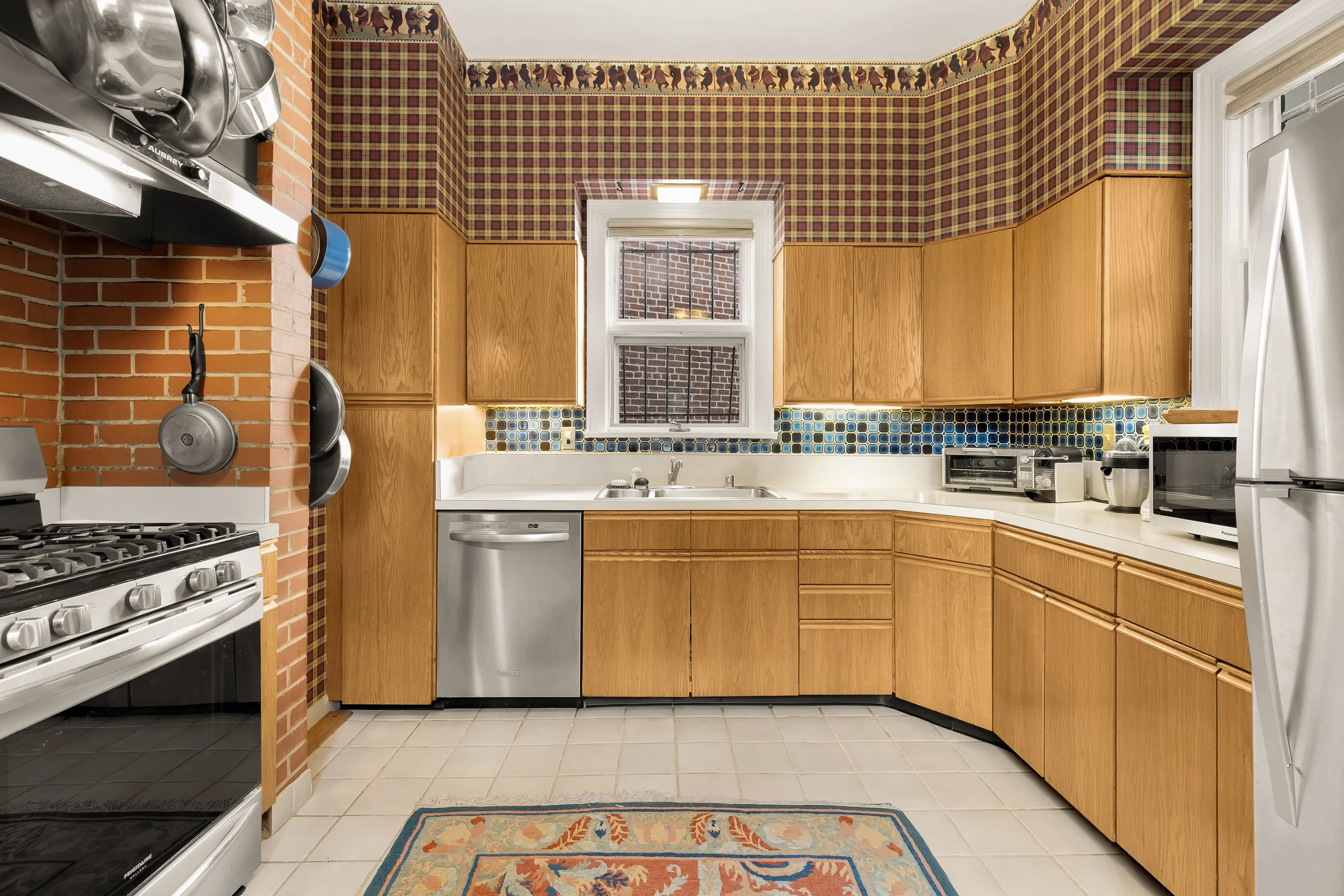 Kitchen with wooden cabinets, brick wall, patterned wallpaper, window, stove, dishwasher, toaster, microwave, and refrigerator.