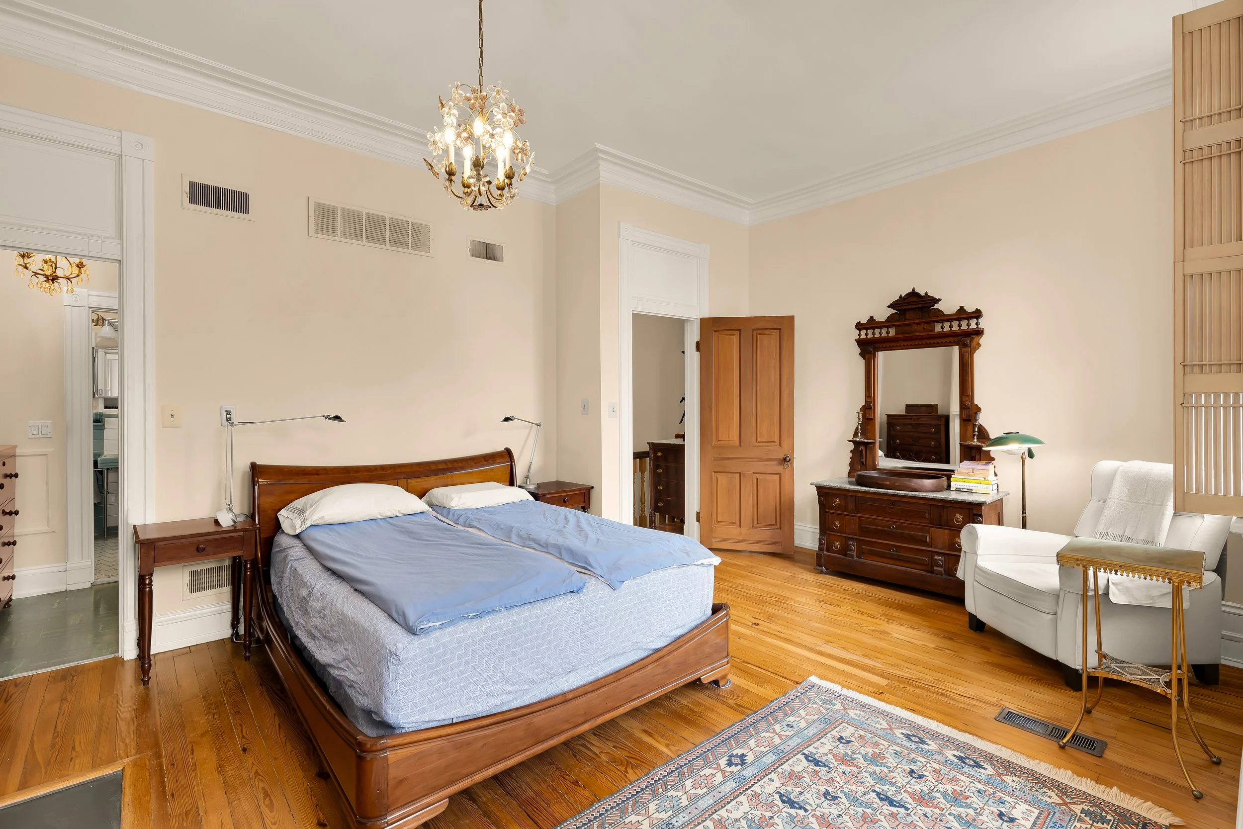 Bedroom with wooden floor, beige walls, antique dresser with mirror, white armchair, and bed with blue bedding.