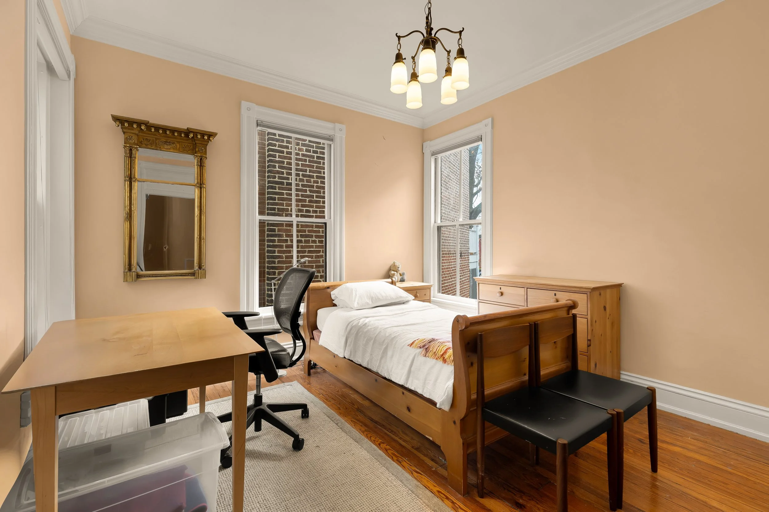 Simple bedroom with wooden bed, side dresser, desk, black office chair, and two black chairs at foot of bed, beige walls, wooden floor, and large windows with brick building outside.
