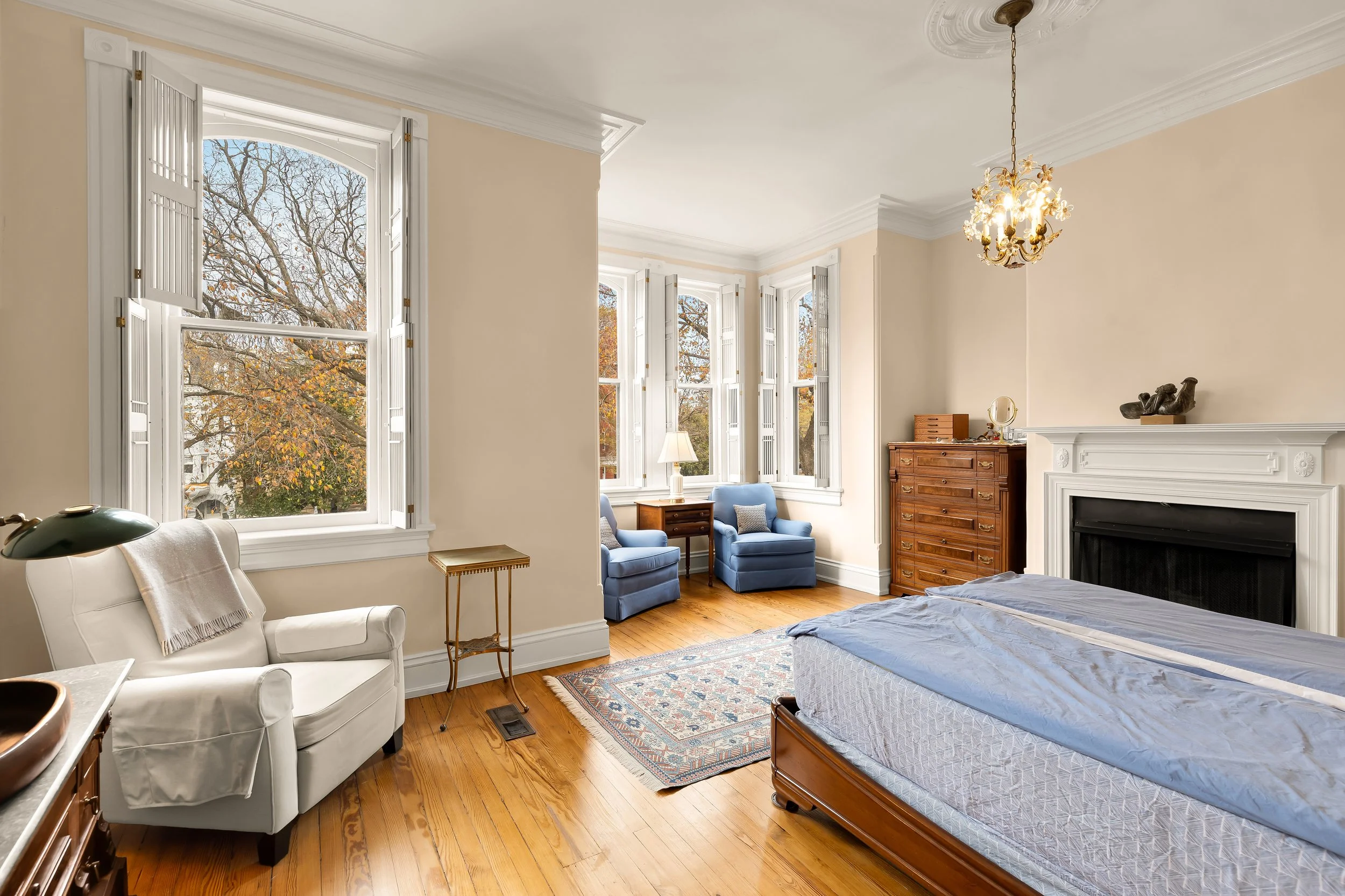 A spacious bedroom with hardwood floors, cream-colored walls, and large windows with white shutters showing trees outside. The room has a white armchair, a small side table, two blue armchairs by the window, a wooden dresser, and a fireplace with dec