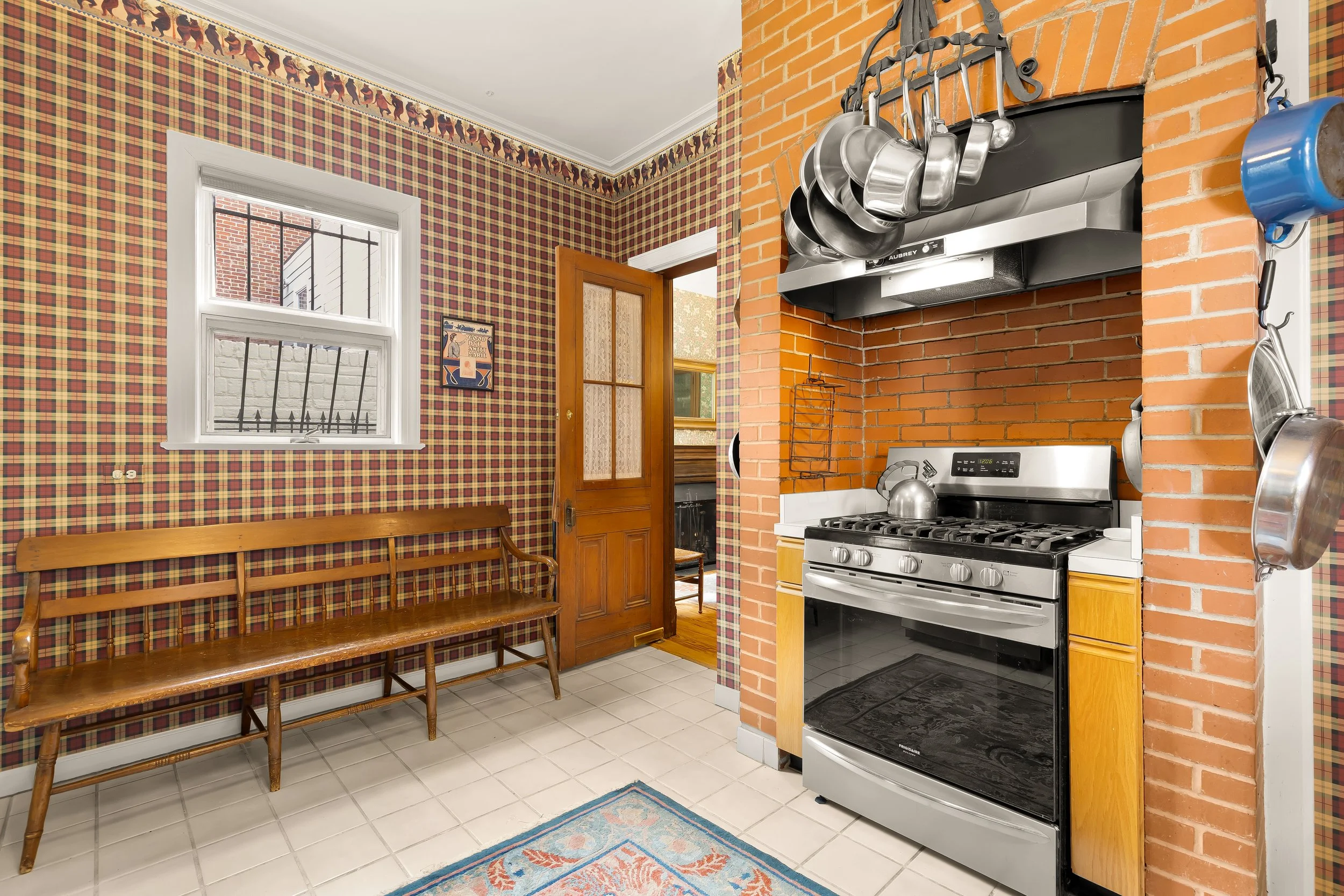 A kitchen with red brick walls and plaid wallpaper. There is a wooden bench, a window with white trim, and a door. The kitchen features a stainless steel stove and oven, with a pot on top. Pots and pans hang from a rack above the stove, and a colorfu