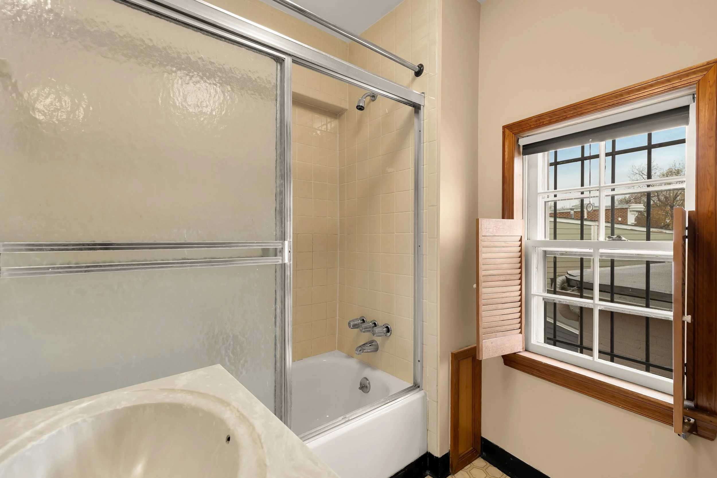 Bathroom with a bathtub and shower, a frosted glass sliding door, a window with wooden trim, and a partially open wooden shutter, outside view of rooftops and trees.