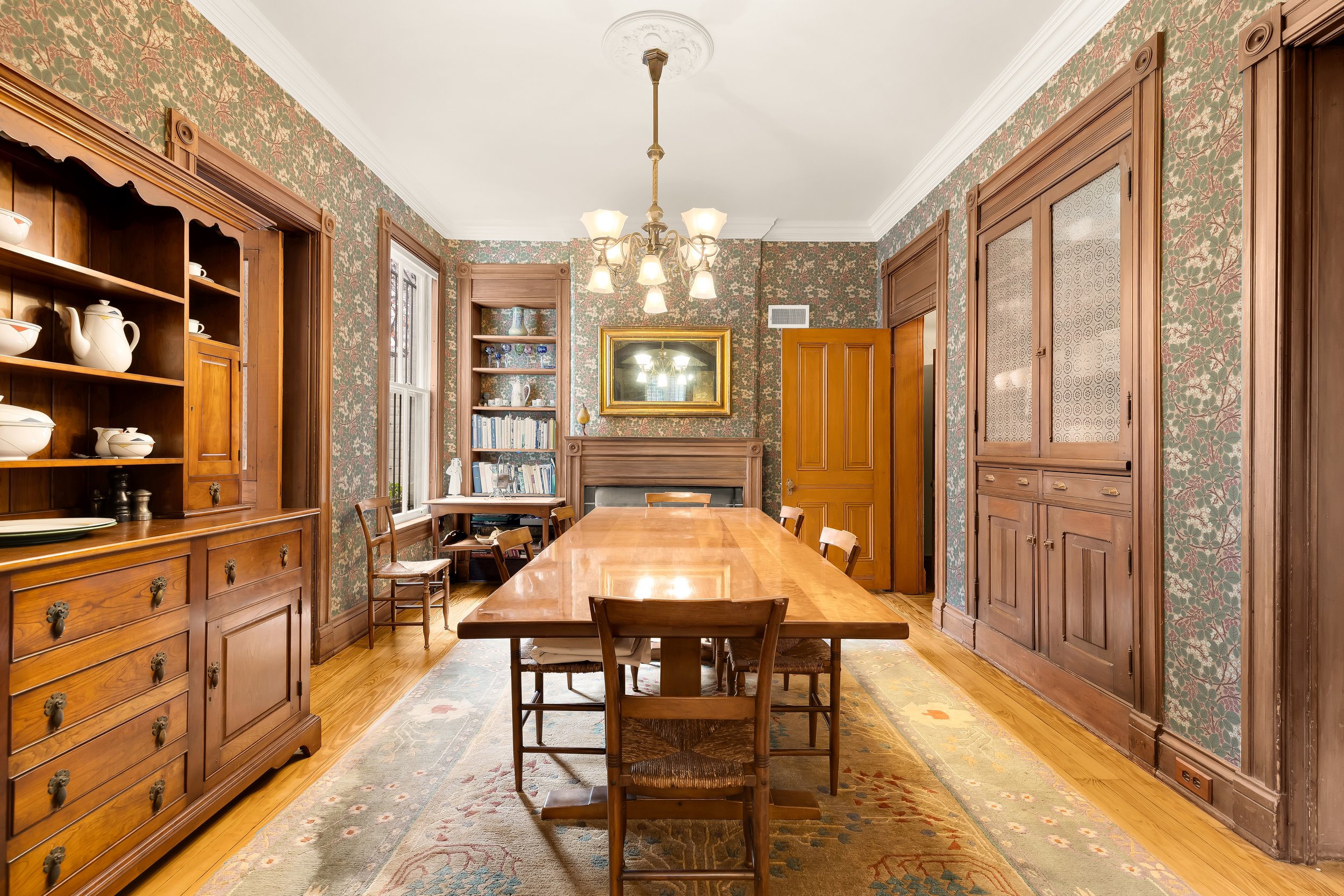 A traditional dining room with wood furniture and floral wallpaper, featuring a large wooden dining table with chairs, a built-in wooden china cabinet, bookshelves, and a chandelier hanging from the ceiling.