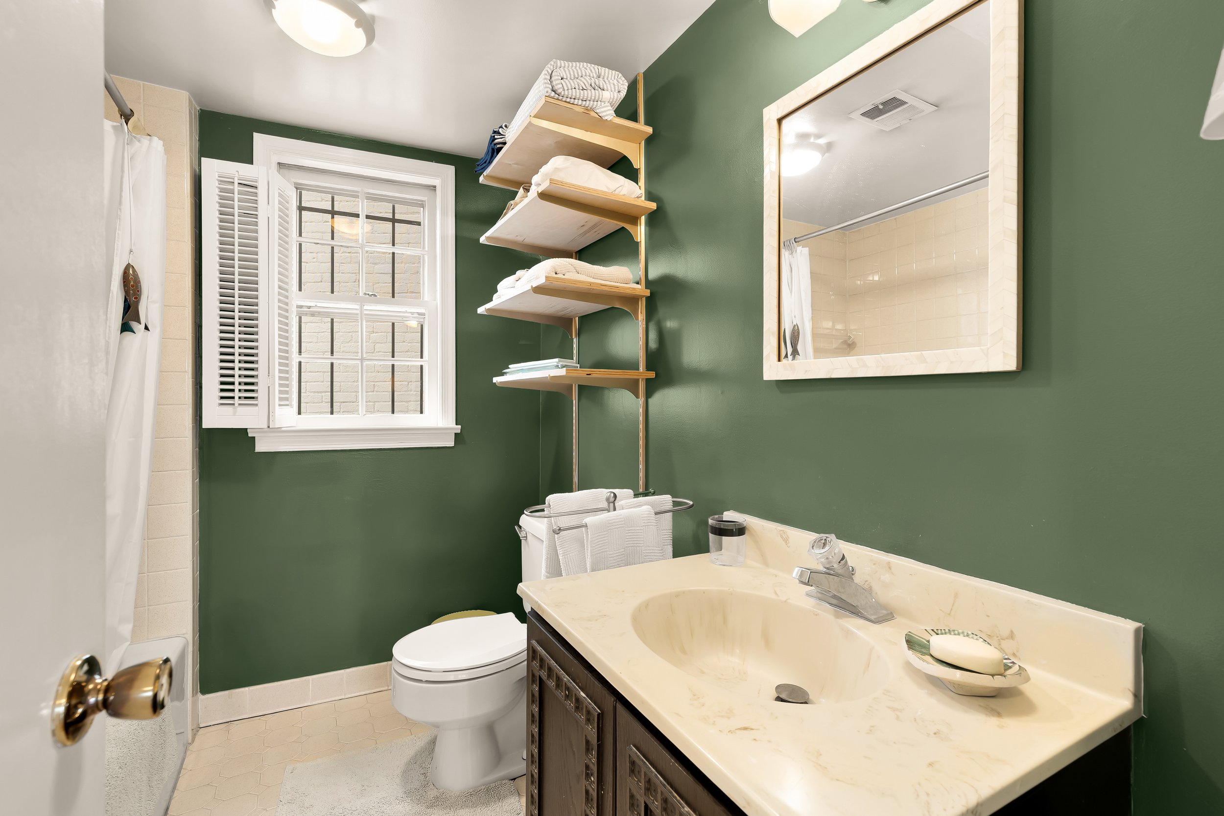 A small bathroom with a green wall, a window with white shutters, a mirror, a ceramic sink with a soap dish, a toilet, and open shelving for towels and linens.