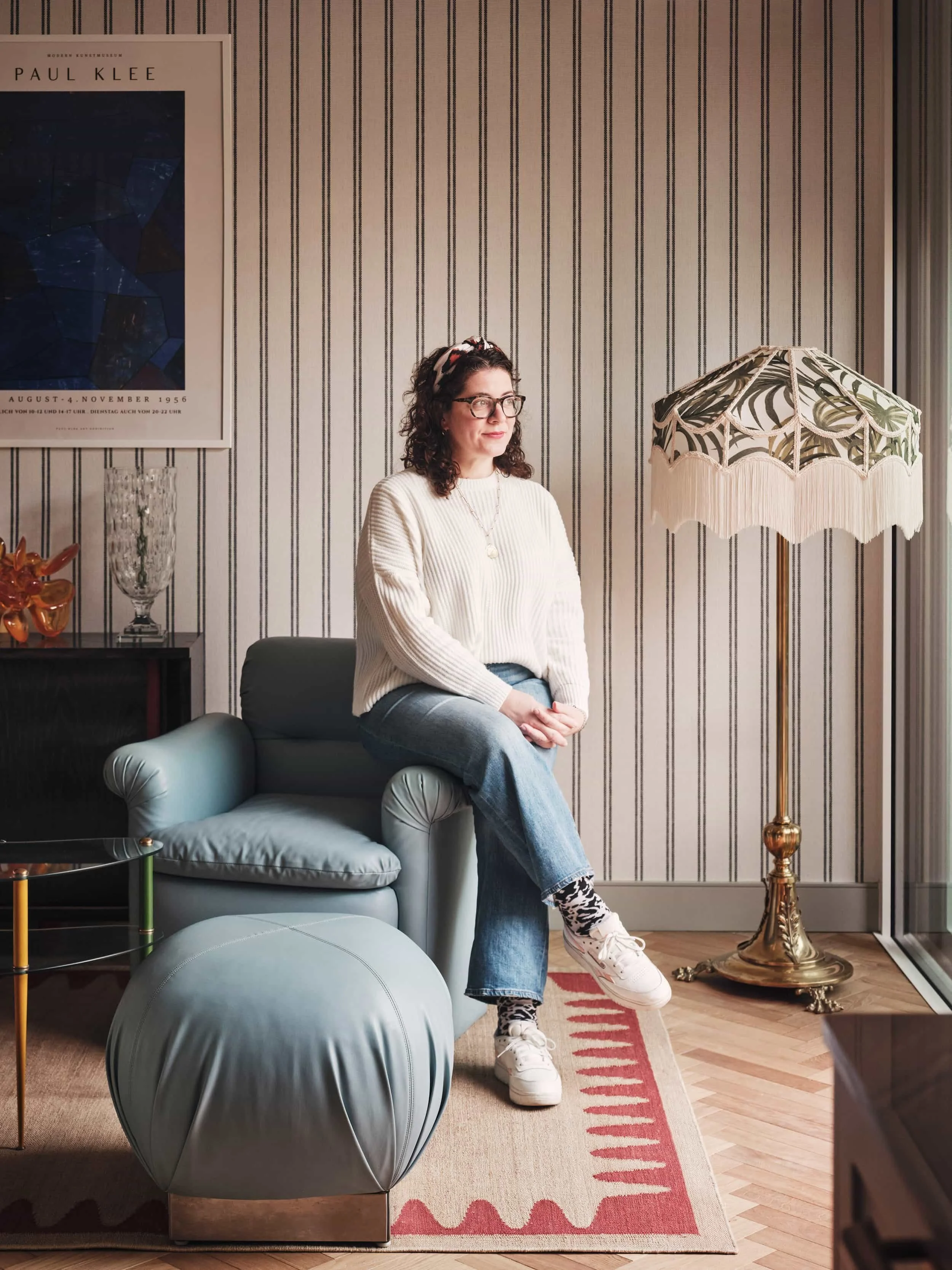 Portrait of Laura Marino seated beside a blue armchair and fringed floor lamp in a Studio L London interior