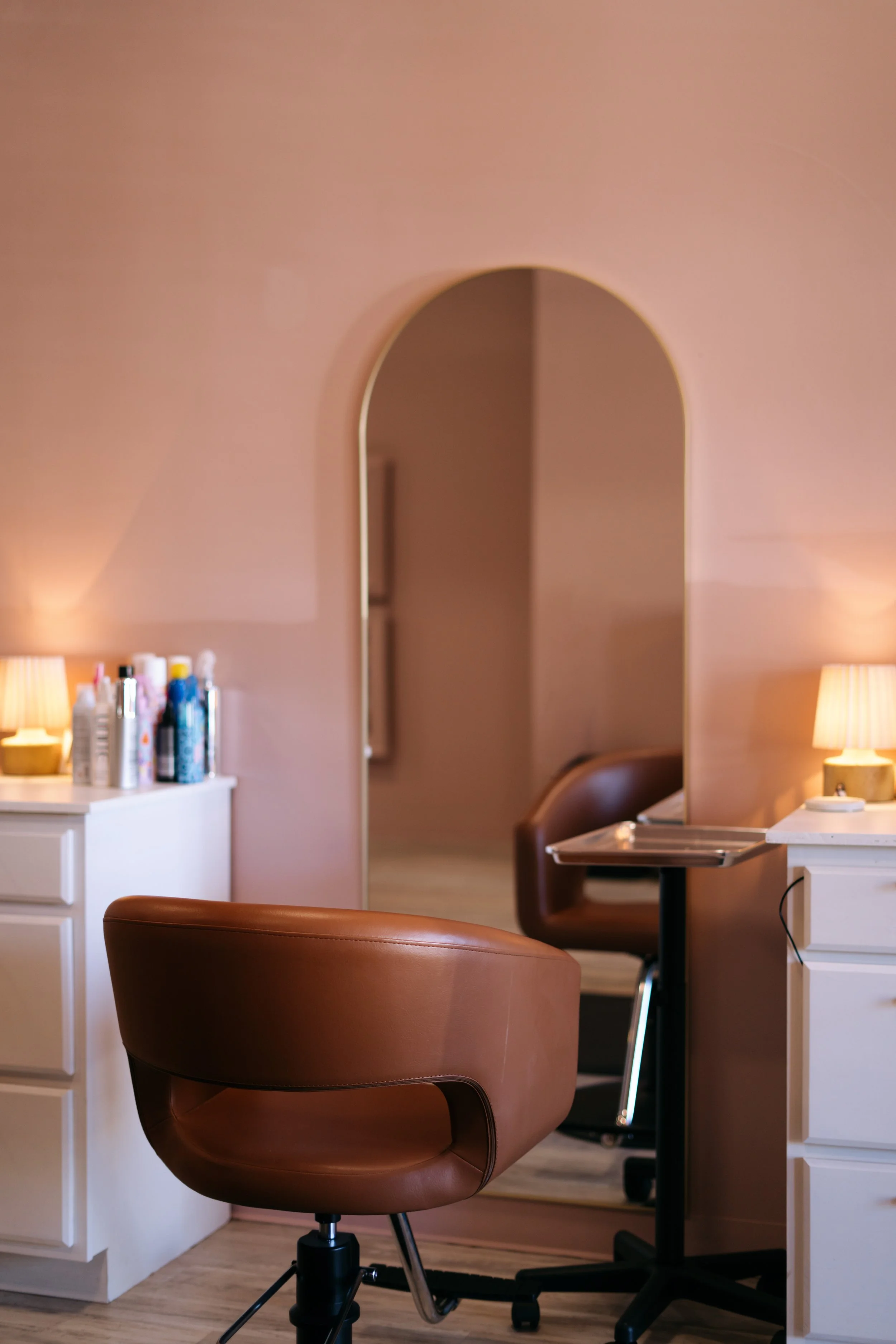 A cozy room with pink walls, featuring a brown leather swivel chair, a white desk with beauty products and hair care bottles, and a corner with a lamp. An arched doorway leads to another part of the space.
