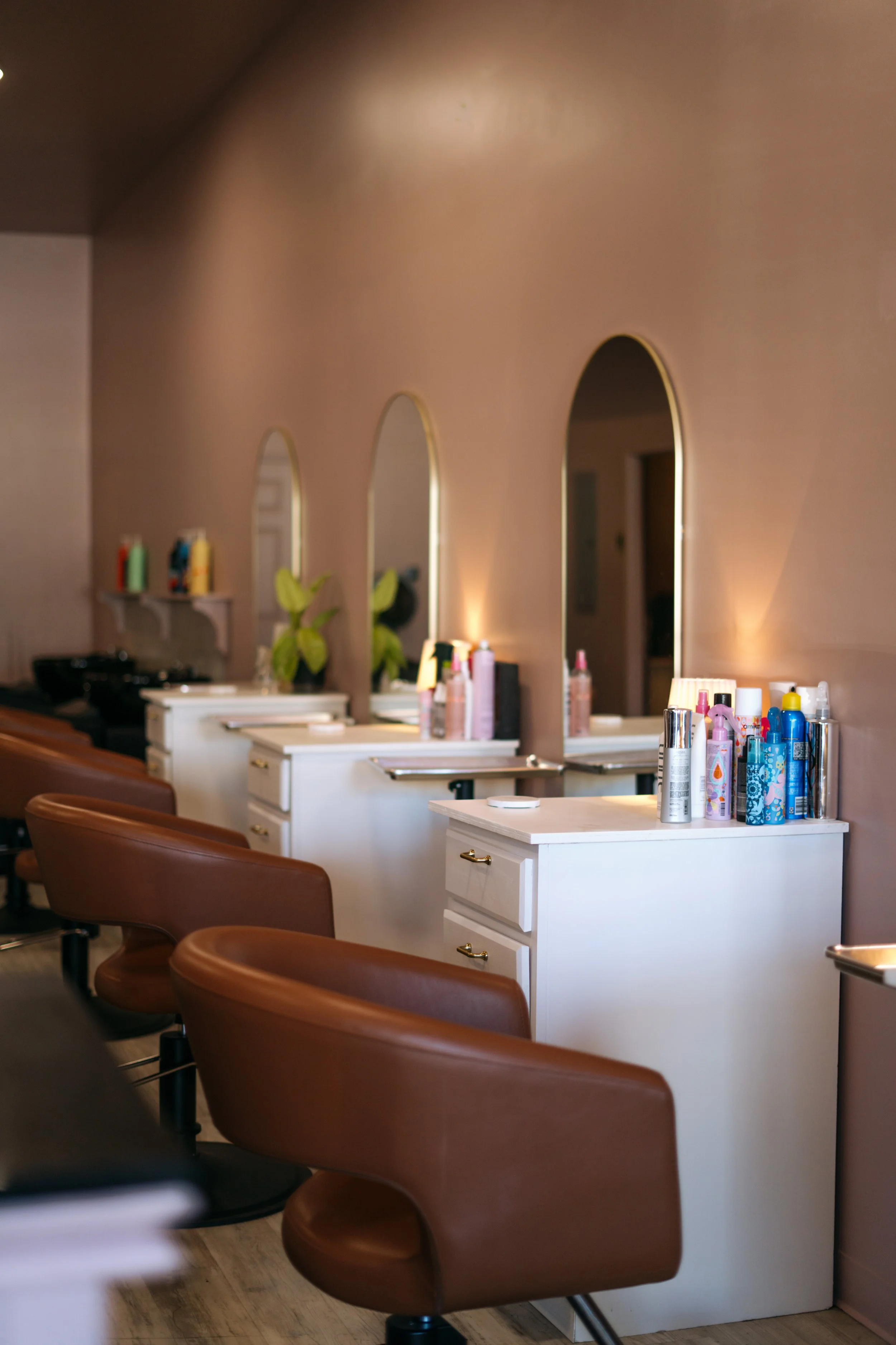 Salon styling stations with white countertops, brown leather chairs, mirrors, and hair care products on the counters.