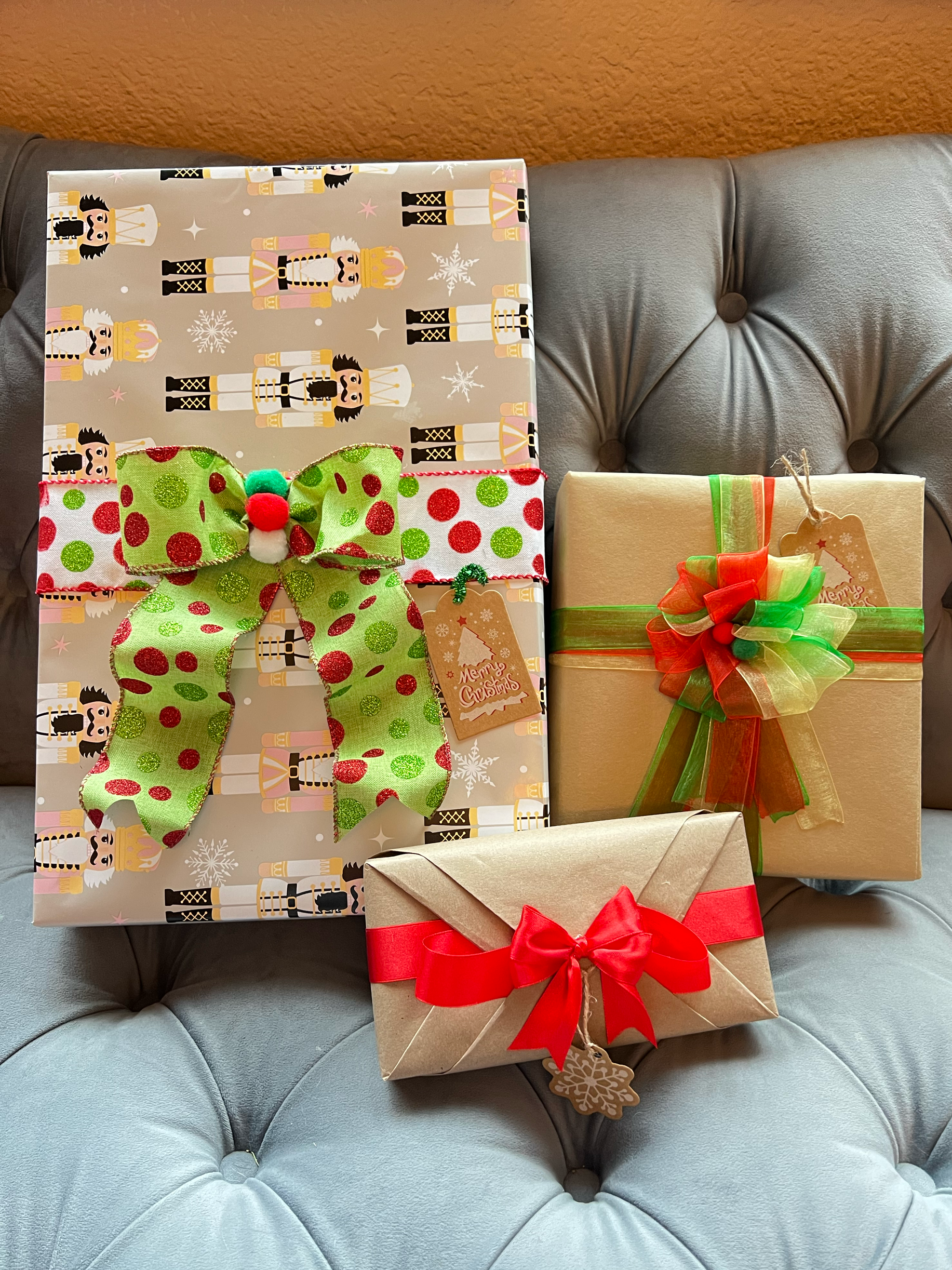 Three wrapped Christmas presents on a gray tufted cushion. The largest is decorated with nutcracker soldier print and a green and red polka dot bow. The second is wrapped in plain beige paper with a large multicolored bow. The third is wrapped in pla