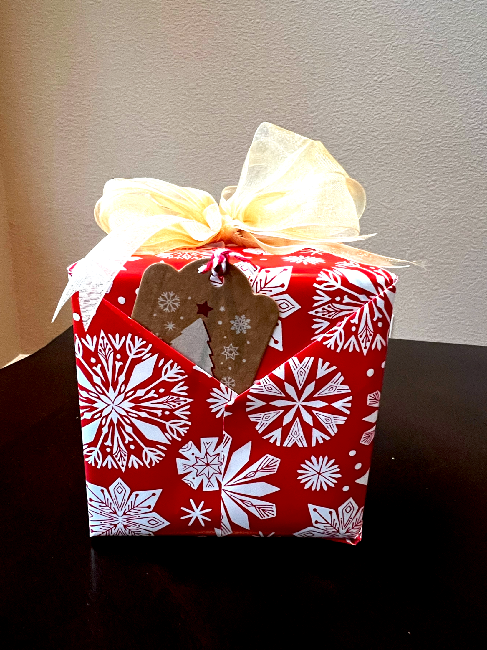 A red Christmas gift box with white snowflake patterns, topped with a cream-colored ribbon bow, and a brown gift tag with a Christmas tree illustration.