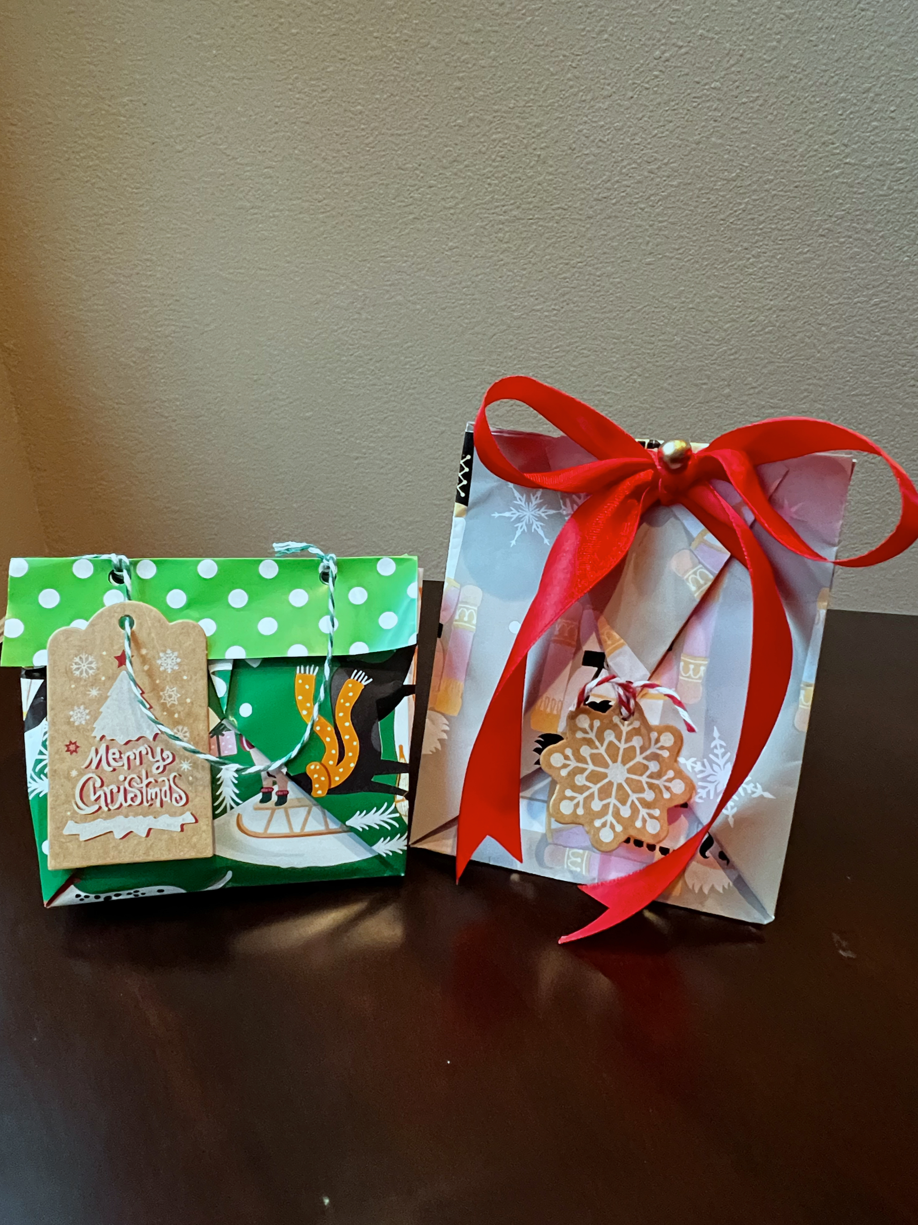 Two decorated Christmas gift bags with holiday tags and ribbons, one green and the other white with red bow, on a dark wooden surface.