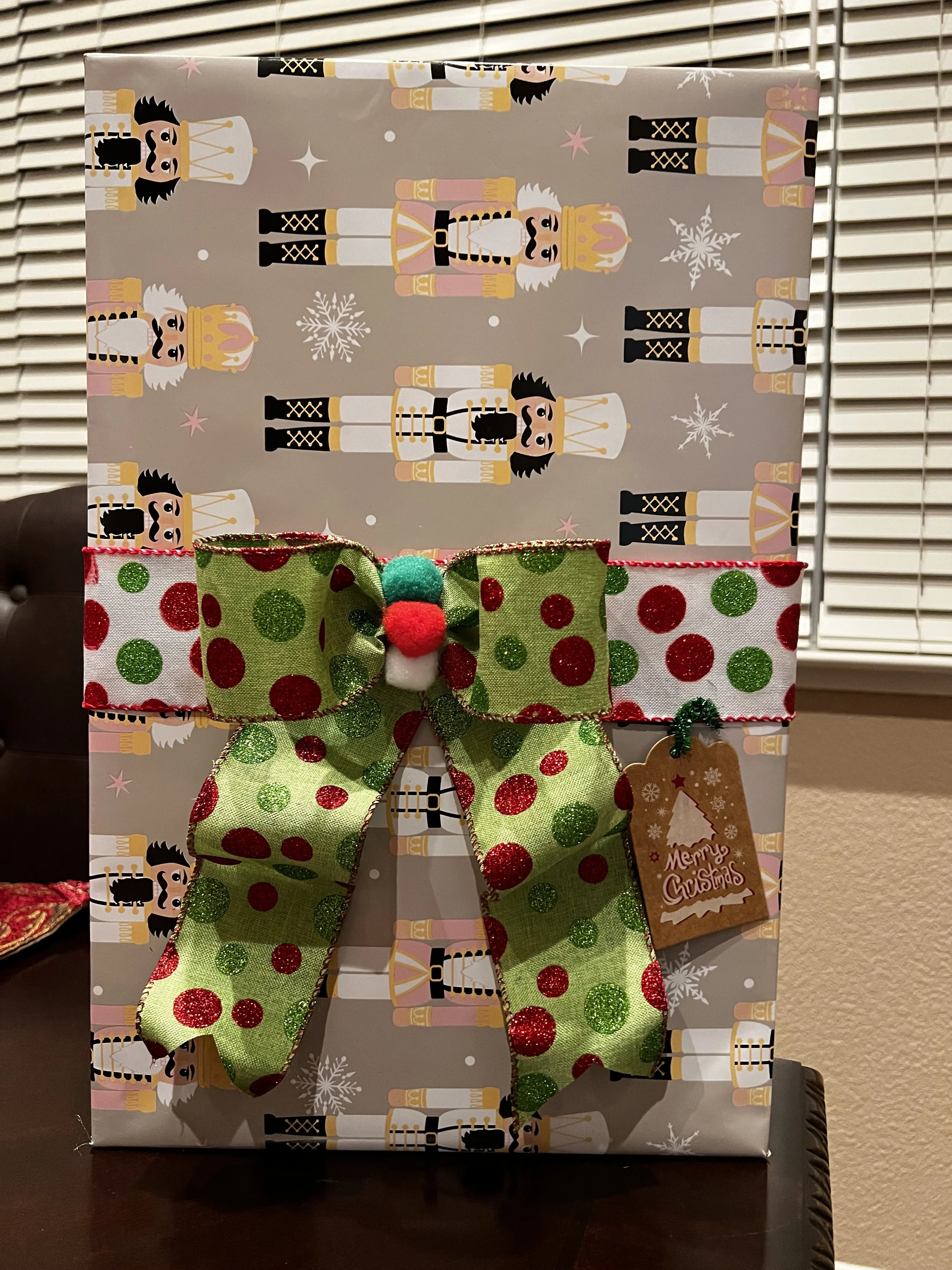 A wrapped Christmas gift with Nutcracker soldier wrapping paper, topped with a green and red polka-dotted bow and a candy-themed tag that says "Merry Christmas."