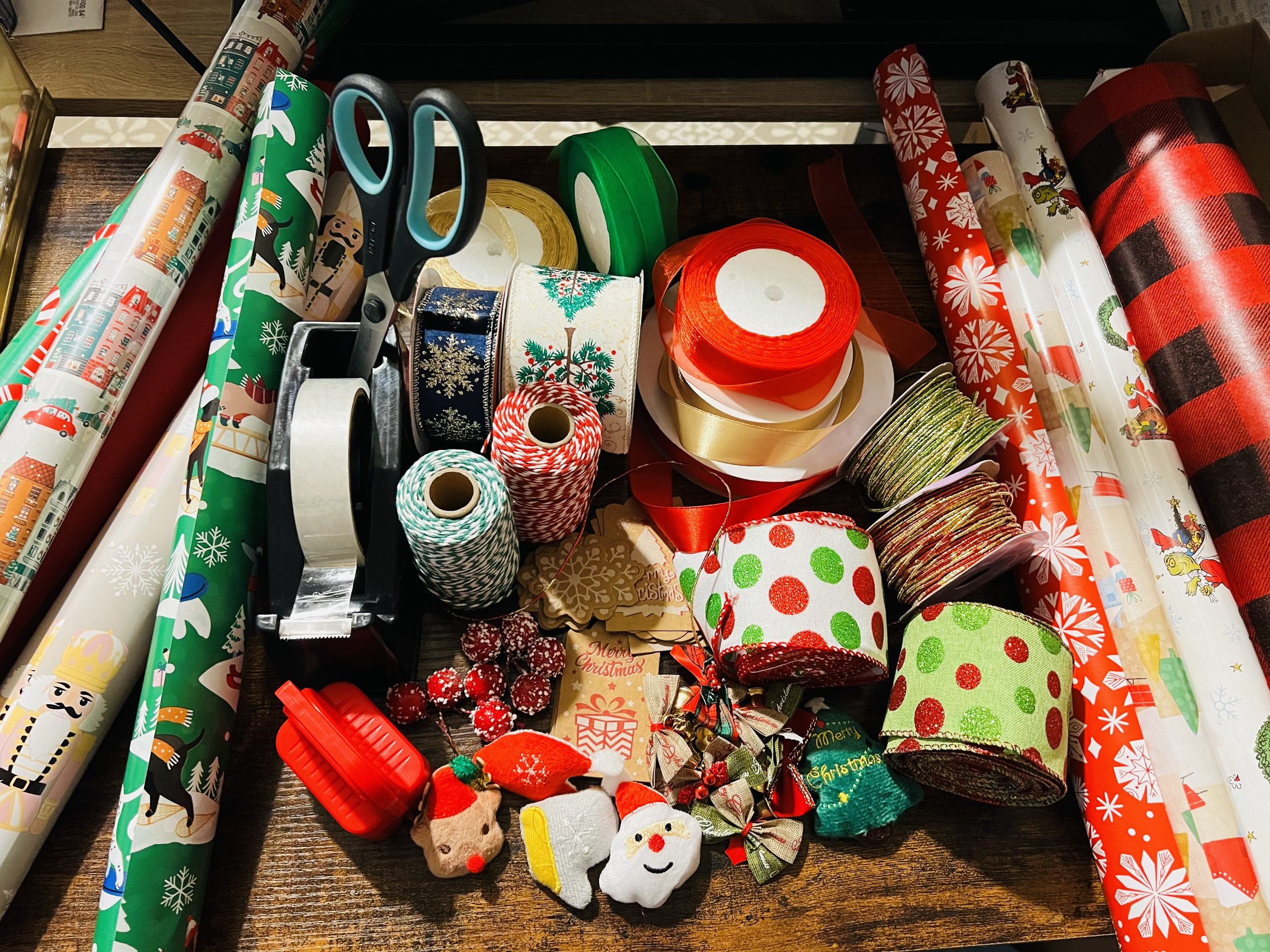 A table with Christmas wrapping paper rolls, scissors, tape, ribbons, ornaments, and small Christmas decorations.