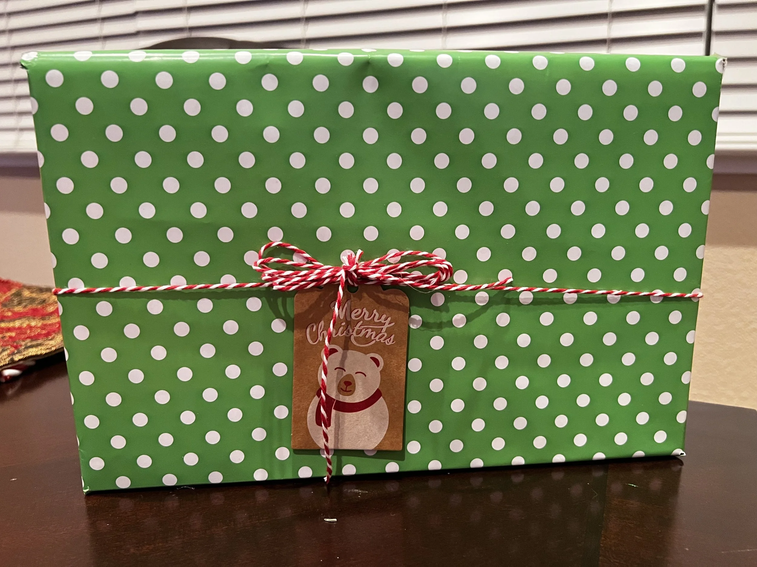 Green gift box with white polka dots, wrapped with red and white twine, and a brown Christmas tag featuring a bear and the text 'Merry Christmas'.