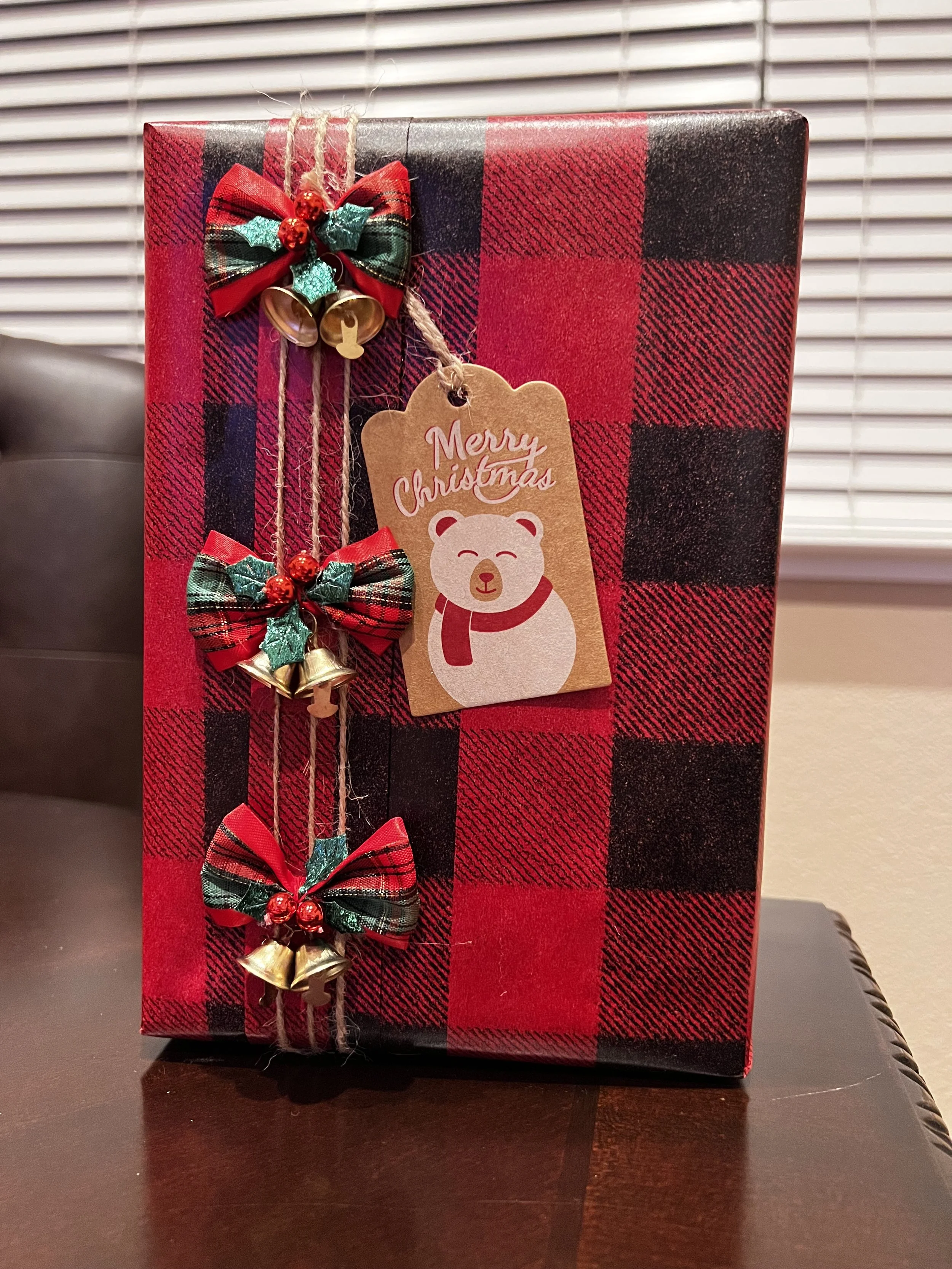 A Christmas gift wrapped in red and black plaid paper with three small holiday bows and bells attached, and a tag with a bear illustration and "Merry Christmas" message.