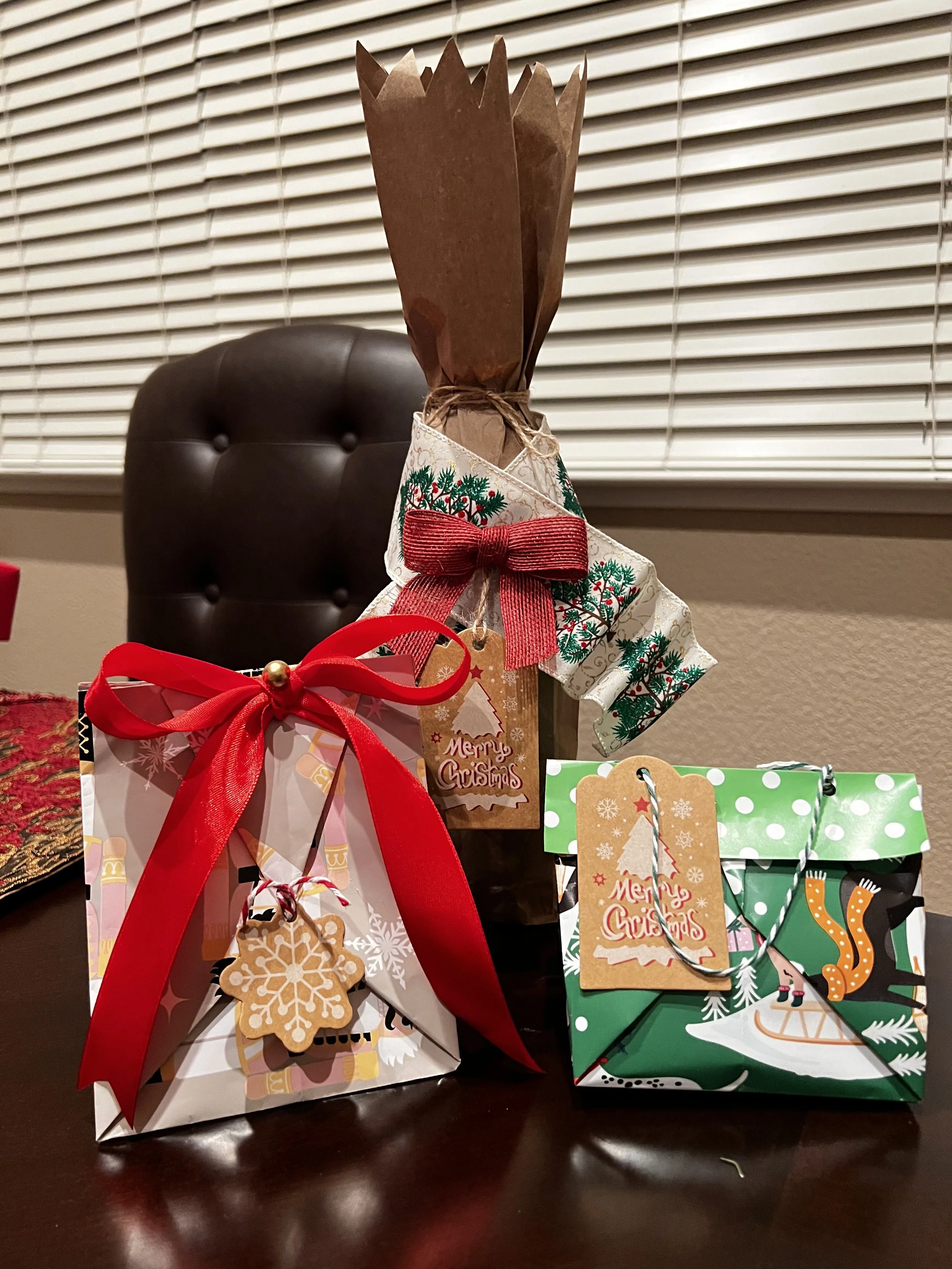 Three Christmas gift packages on a dark wooden table, one wrapped in white paper with a red ribbon and a gingerbread cookie ornament, another in green paper with a Christmas scene and a hanging tag, and the third decorated with Christmas-themed cloth