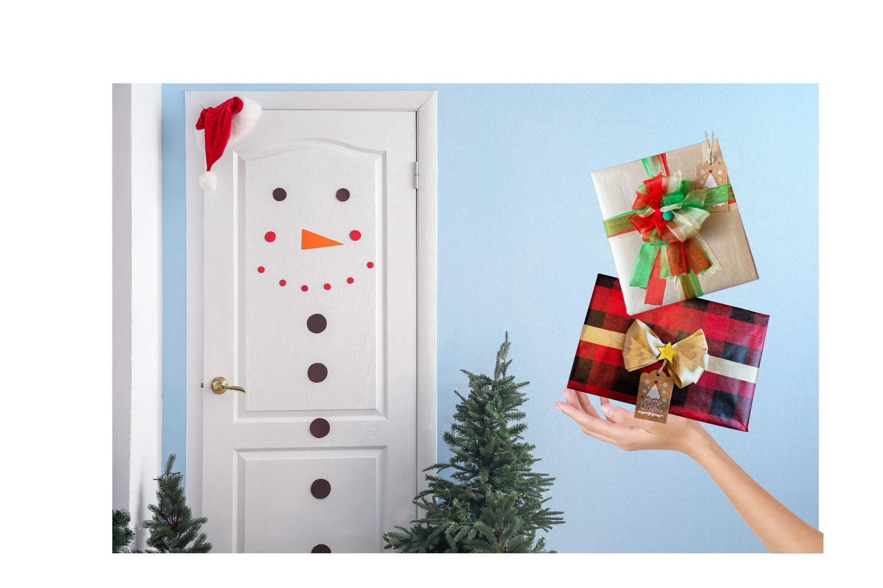 Decorated door with a snowman face made of paper and a Santa hat, small Christmas trees, and a hand holding two wrapped Christmas presents against a light blue wall.