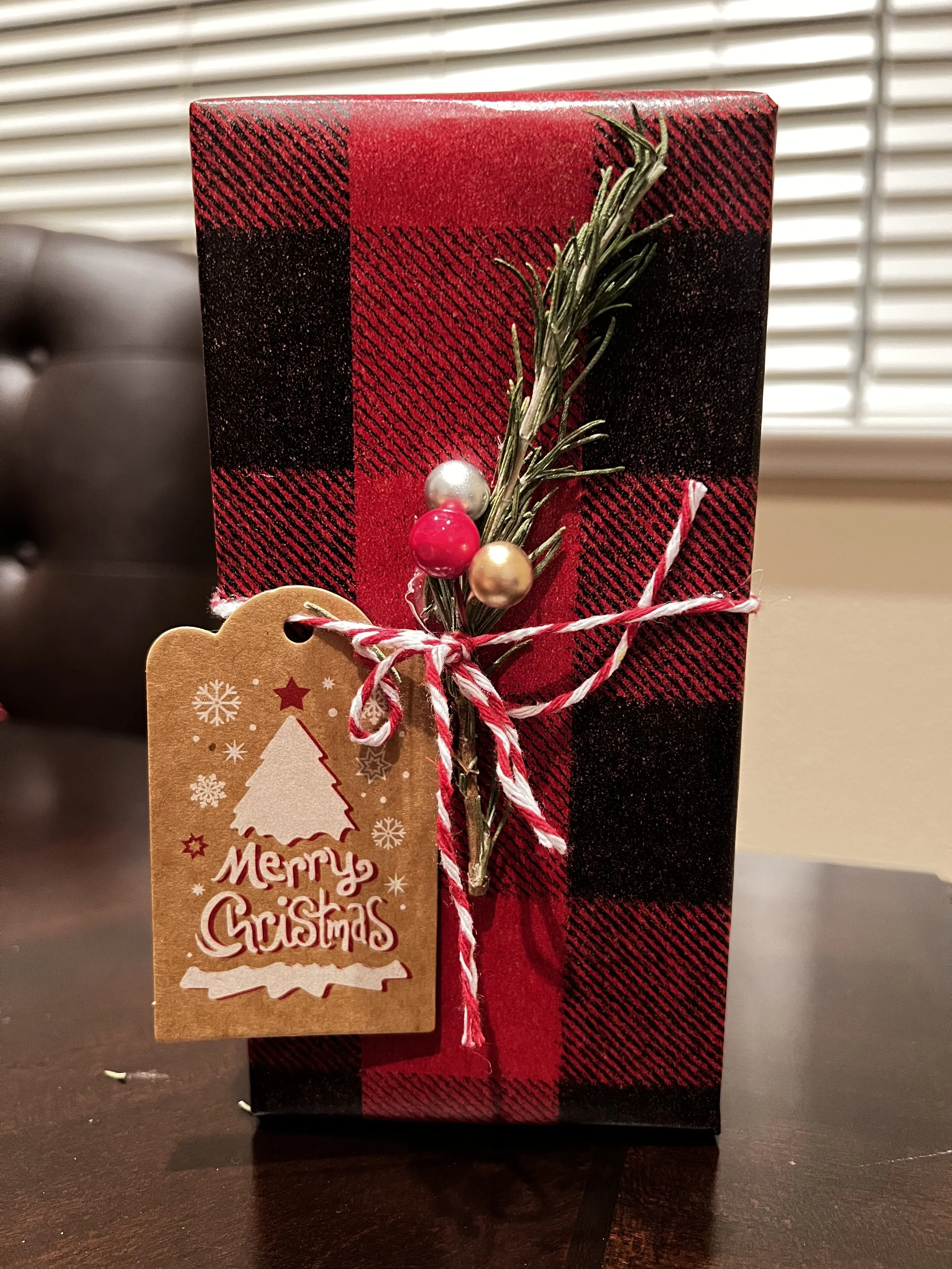 A wrapped Christmas gift in red and black plaid paper decorated with a sprig of greenery, colorful berries, and a 'Merry Christmas' tag.