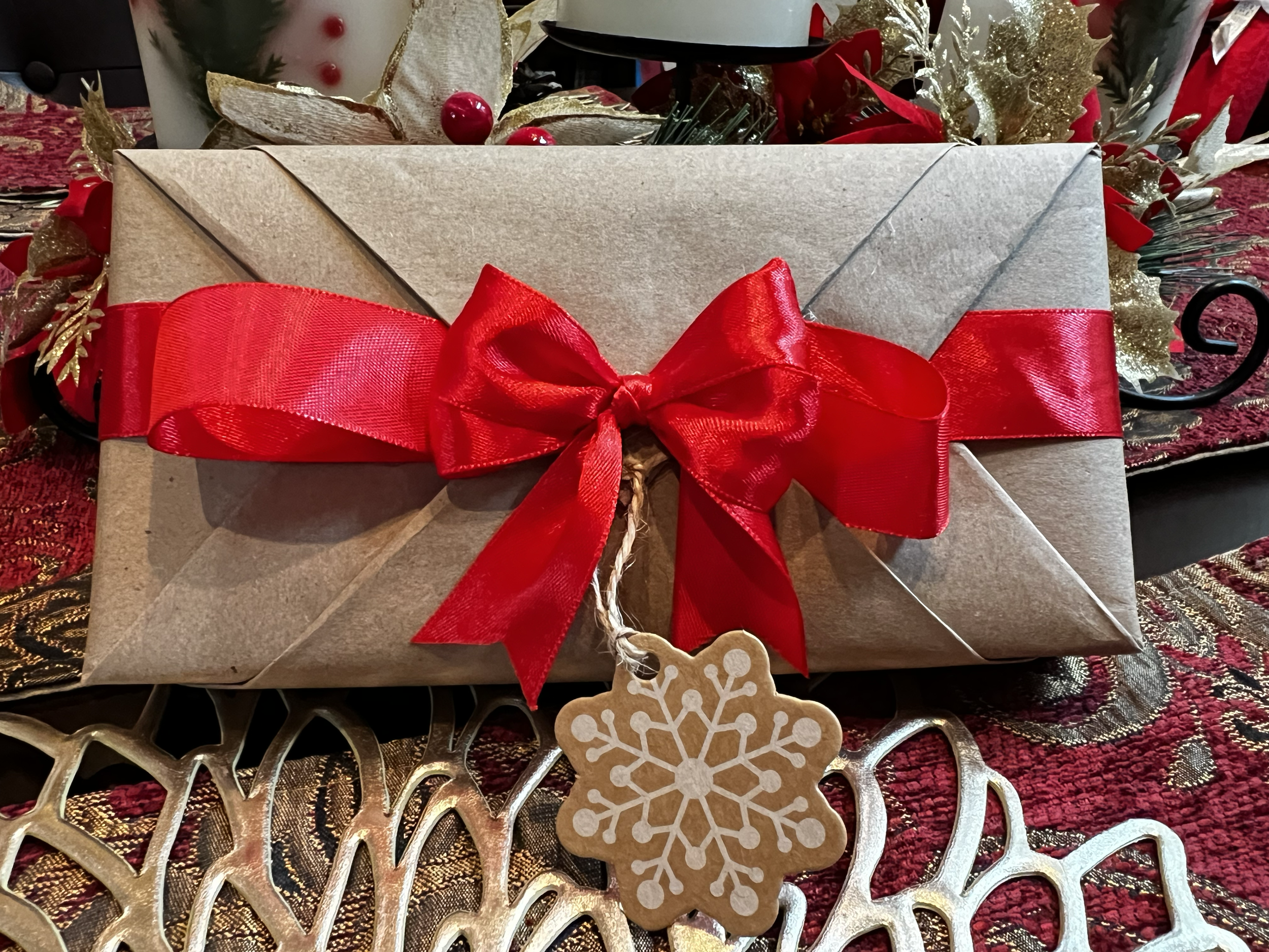 A Christmas gift wrapped in brown paper with a big red ribbon and bow, adorned with a snowflake ornament, placed on a festive tablecloth surrounded by holiday decorations.
