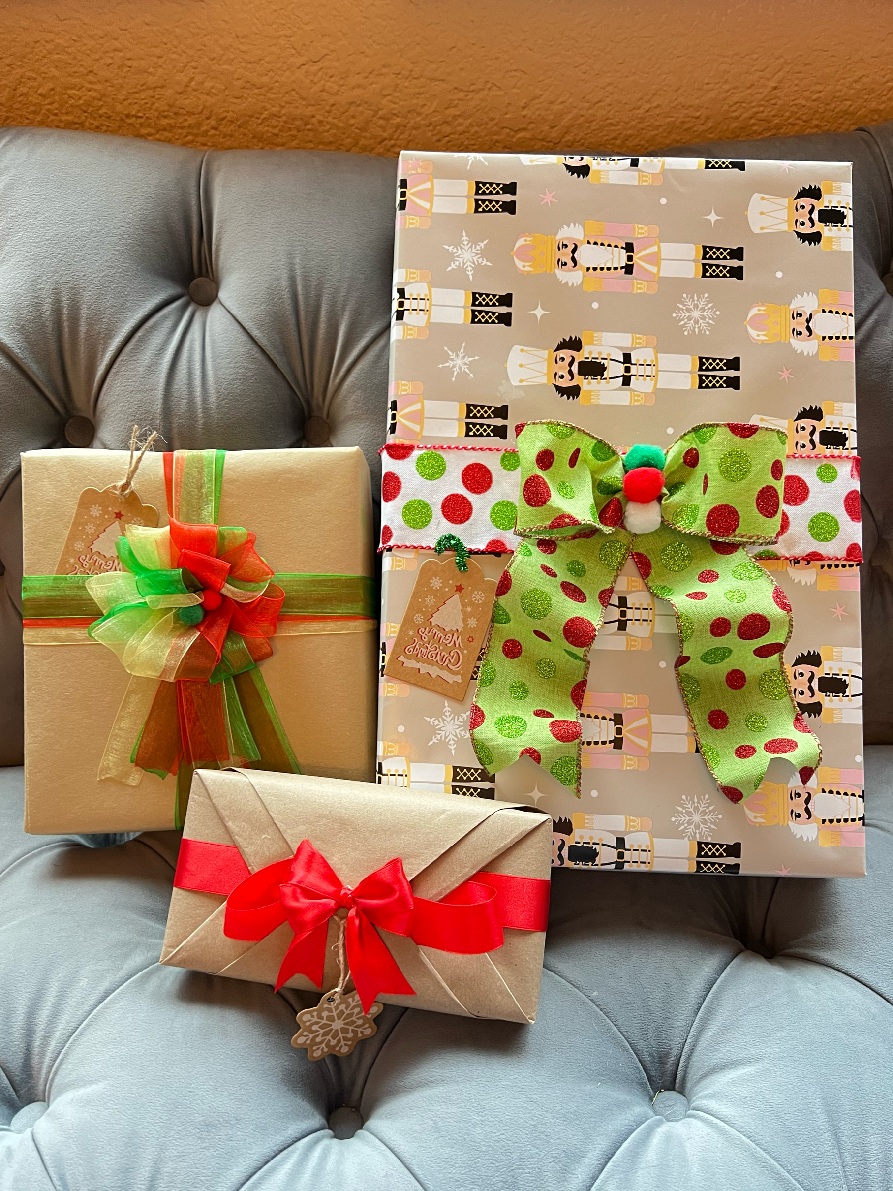 Three wrapped Christmas presents placed on a gray, tufted, leather couch with a brown wall in the background. The largest gift has holiday-themed wrapping paper with toy soldiers and snowflakes, and a large green and red polka-dot bow with a red pom-pom. The other two gifts are wrapped in plain beige paper with decorative bows, one with a green and red bow with a small tag, and the smaller with a red ribbon and a snowflake tag.