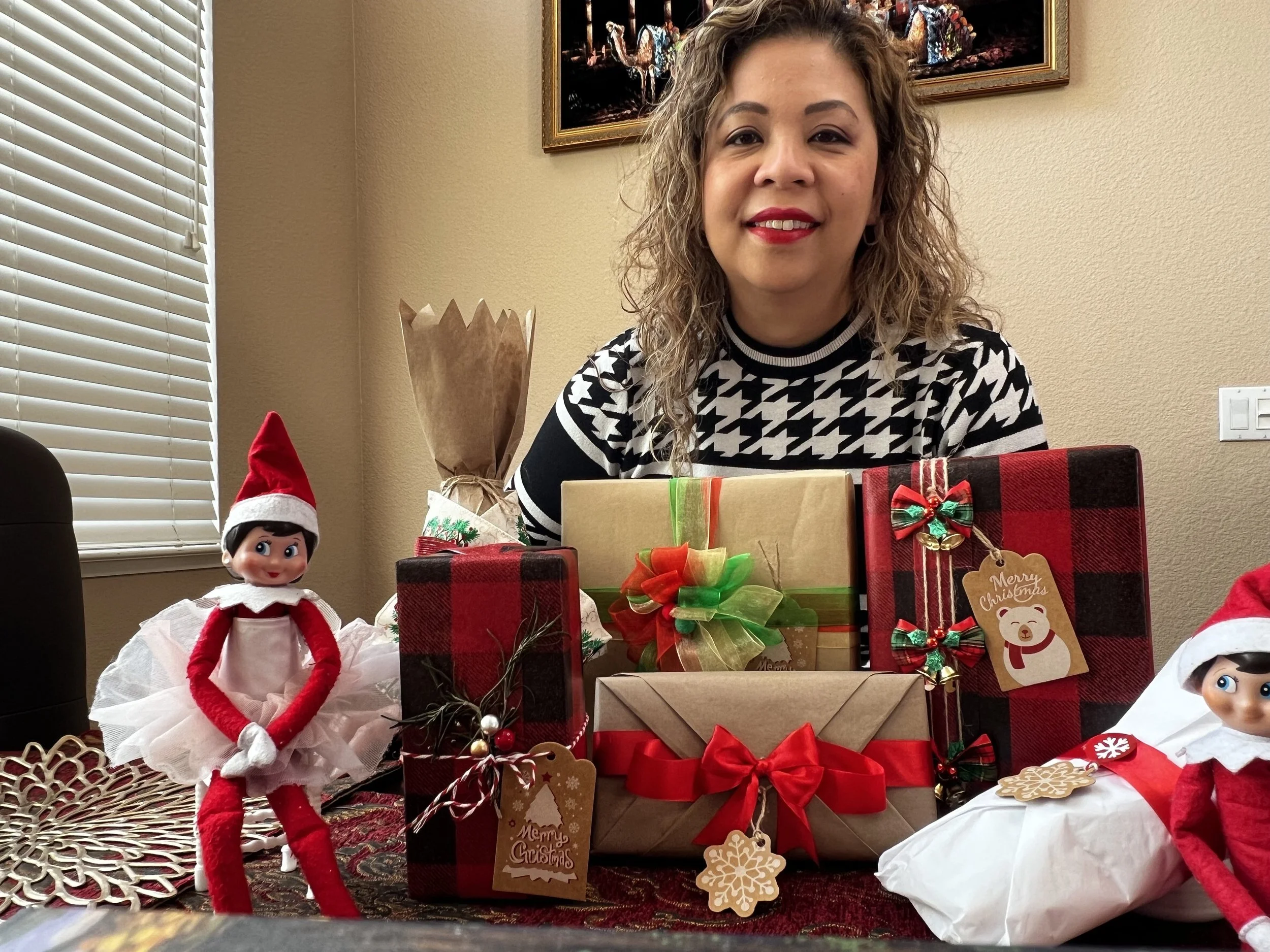 A woman with curly hair and red lipstick sitting behind a table with Christmas gifts and decorations, including plush Santa and elf dolls, wrapped presents, and holiday tags, in a room with beige walls and a painting on the wall.