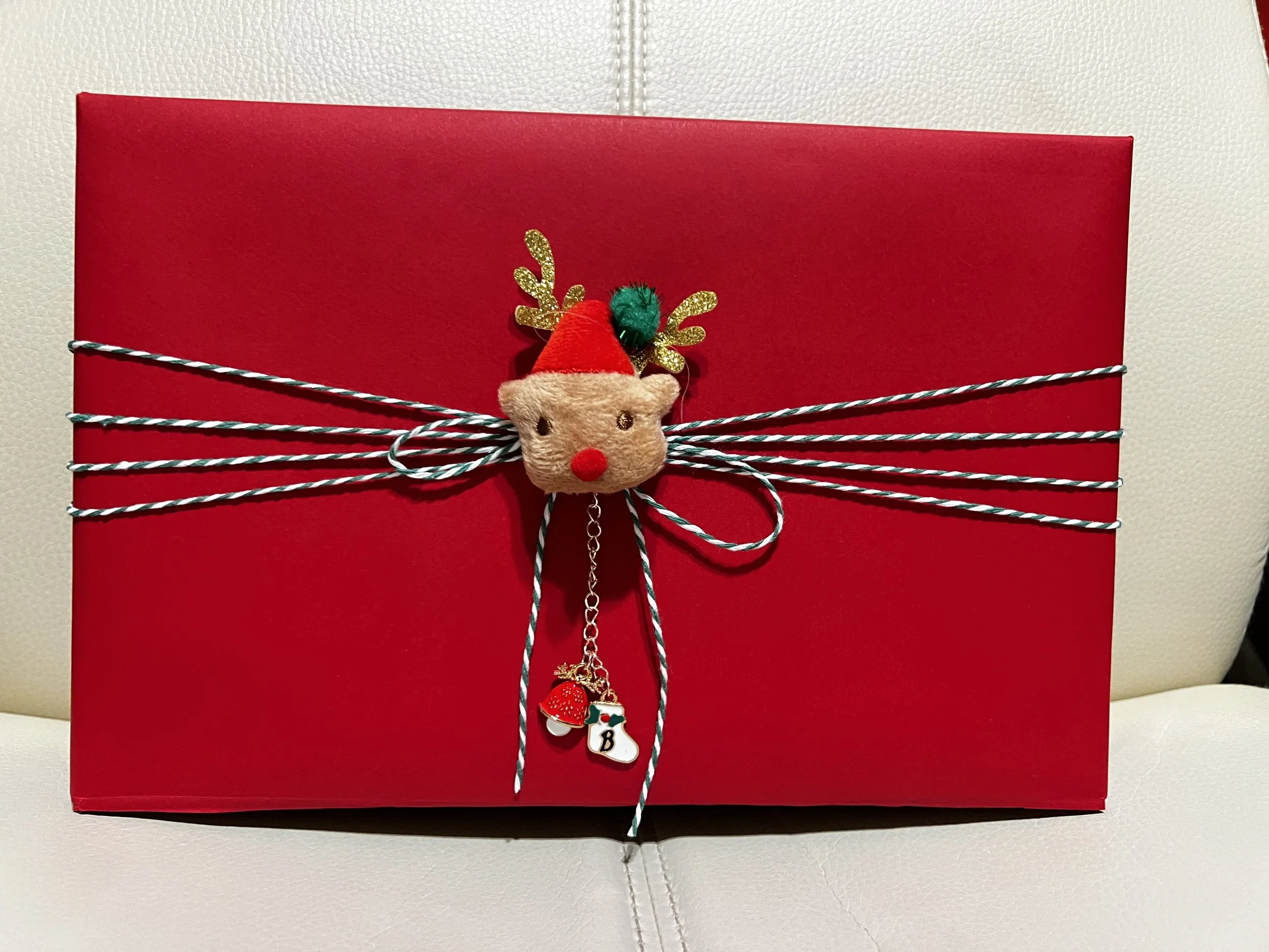 Red Christmas gift box decorated with a reindeer face ornament, a red pom-pom nose, gold reindeer antlers, and a red Santa hat, tied with red and white striped twine and holiday charms.