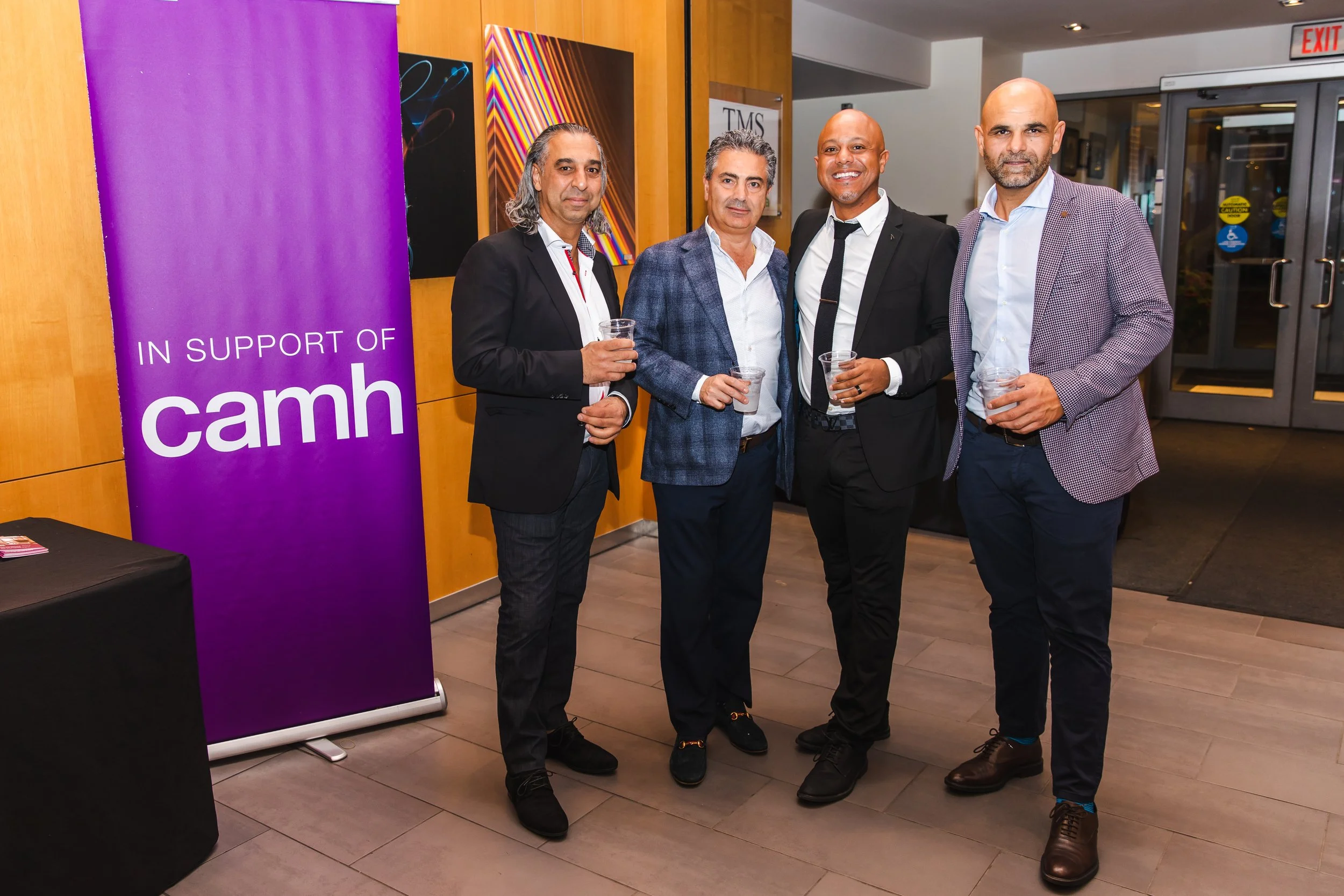 Four men in suits standing indoors at an event with a purple banner that reads 'In Support of CAMH' on the left side of the image. They are holding drinks and smiling at the camera.