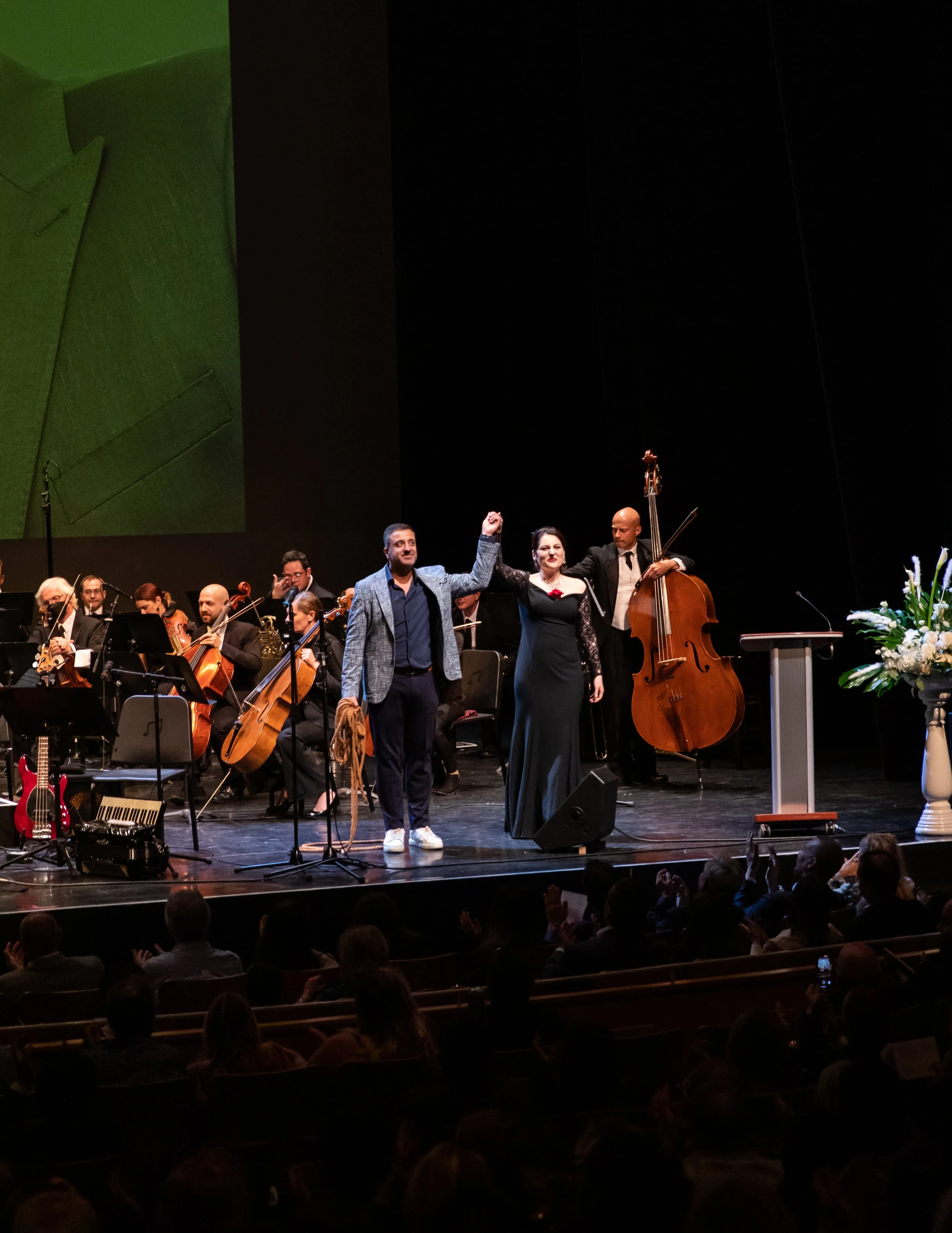 On stage, a man and a woman are holding hands and raising their fists in triumph, with an orchestra seated behind them. The audience is clapping.