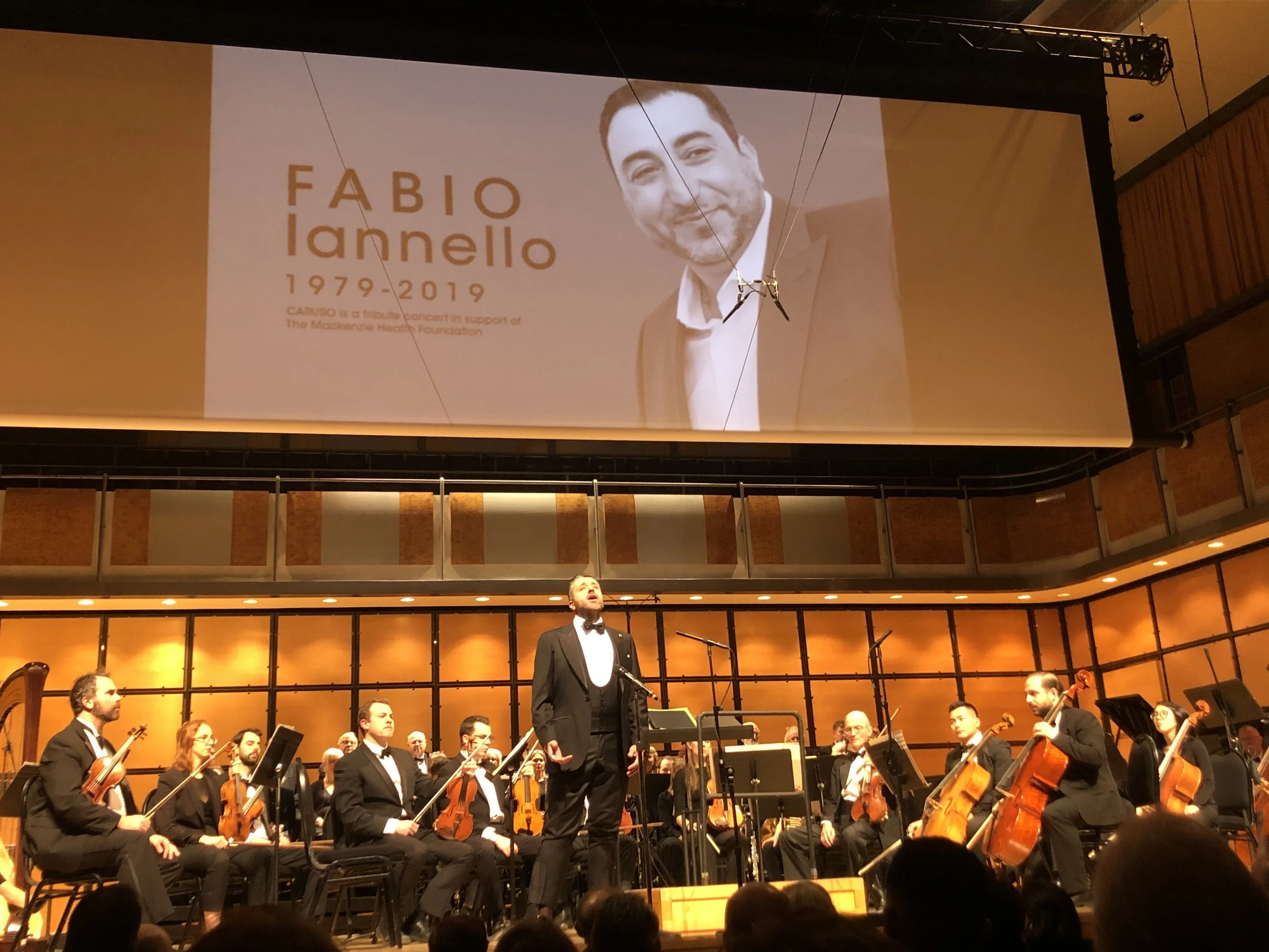 A formal symphony orchestra performing on stage with a conductor in a tuxedo, and a large screen above displaying a tribute to Fabio Iannello, with a photo of him, his name, and dates 1979-2019.