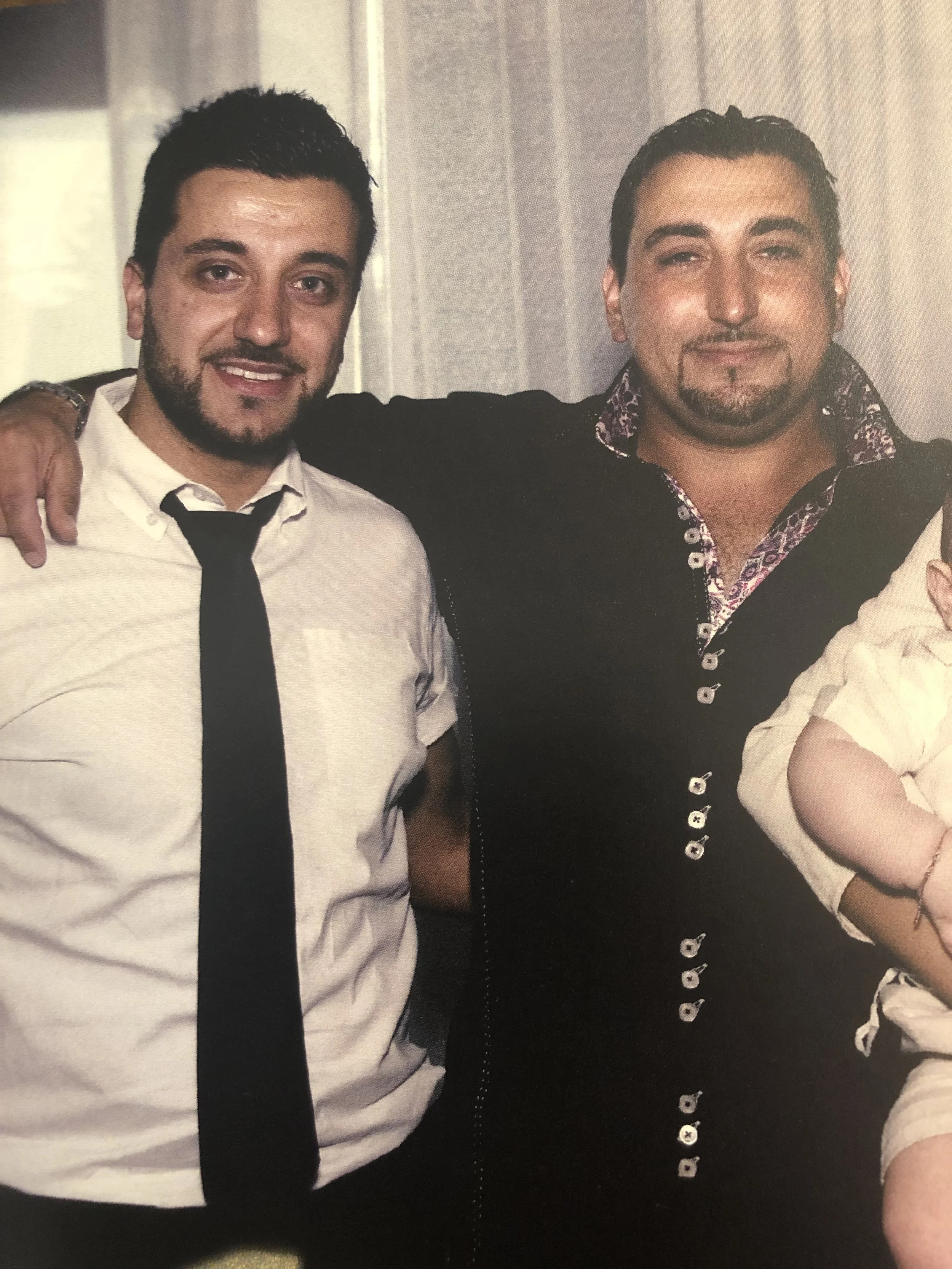 Two men smiling, one with arm around the other, at a social gathering or event.