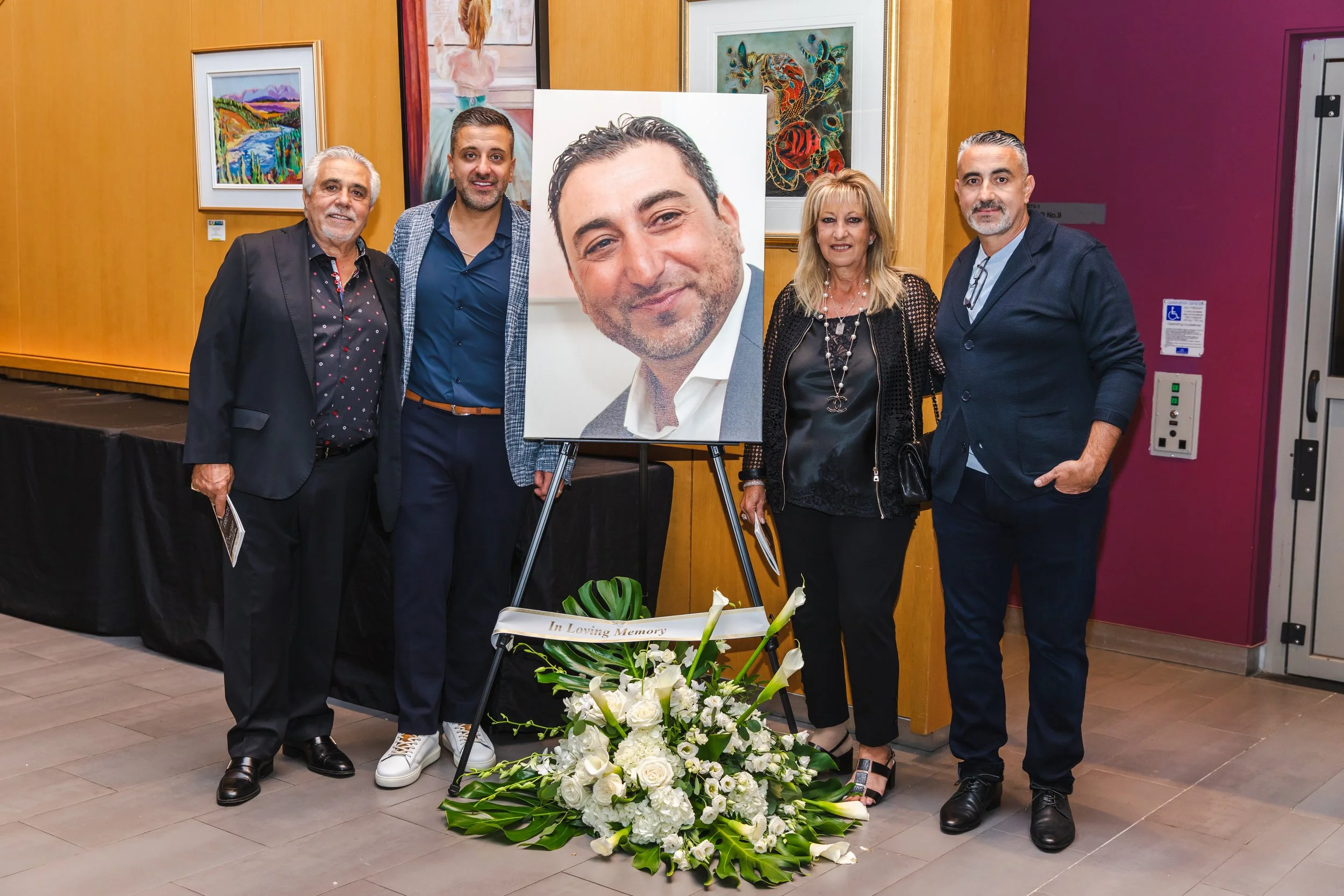 Group of five people standing together at a memorial event, with a large portrait of a man's face and a floral arrangement in front. The setting appears to be an art gallery or event space.