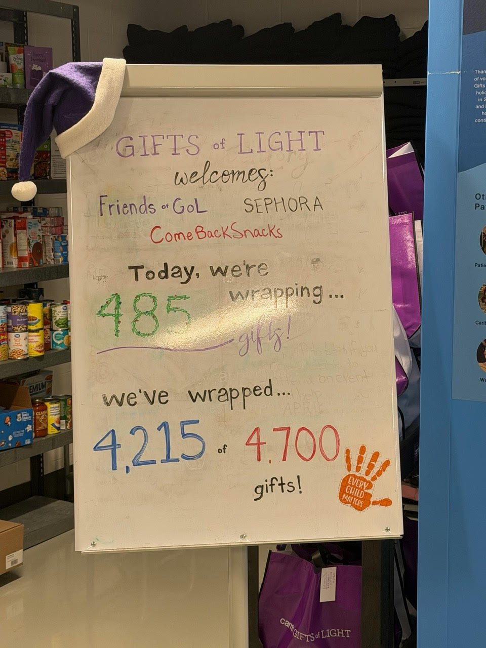 Whiteboard with handwritten message about gift wrapping, numbers indicating gifts wrapped and to be wrapped, decorated with a purple Santa hat with white trim and pompom, and an orange handprint sticker that says 'Every Child Matters'.