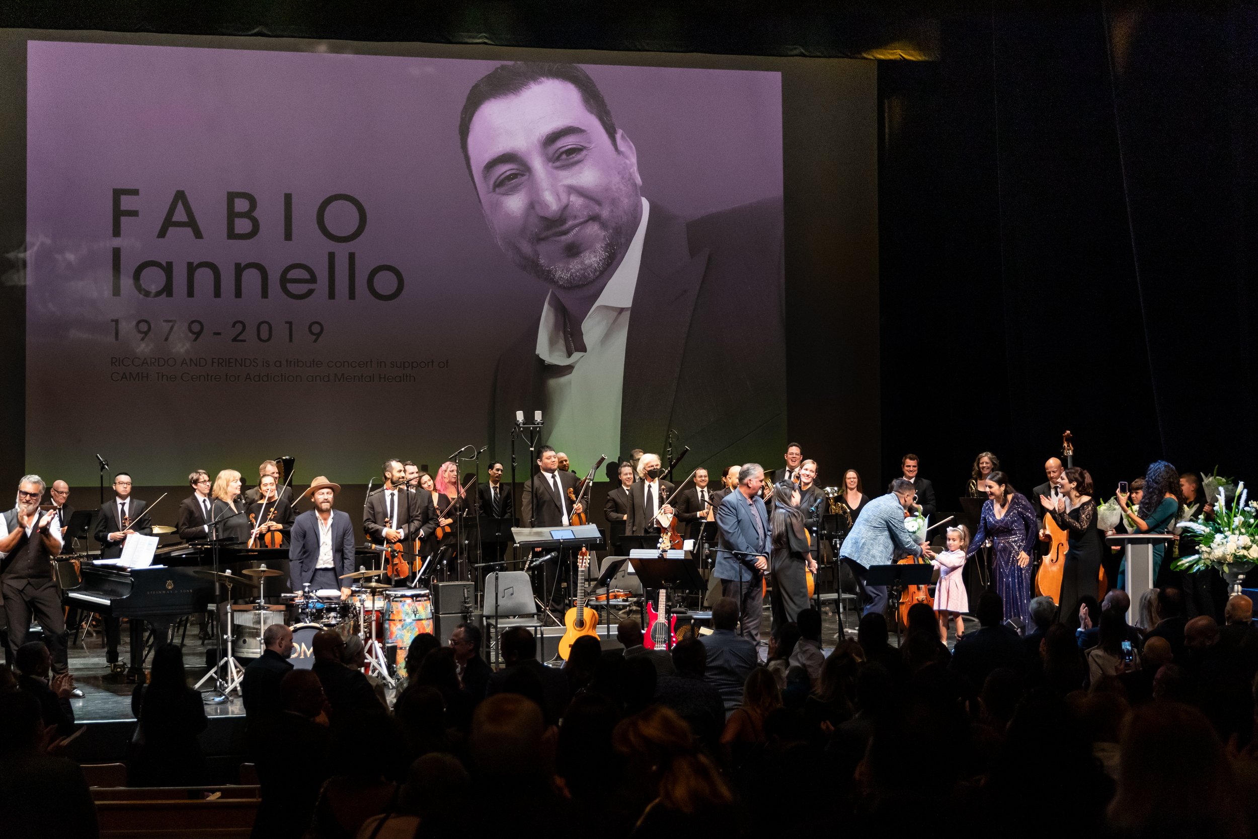 A stage with a large screen displaying a tribute to Fabio Iannello, featuring his photo and dates 1979-2019. The stage is filled with musicians, some holding instruments. A man in a gray suit is presenting flowers to a woman in a purple dress with a 