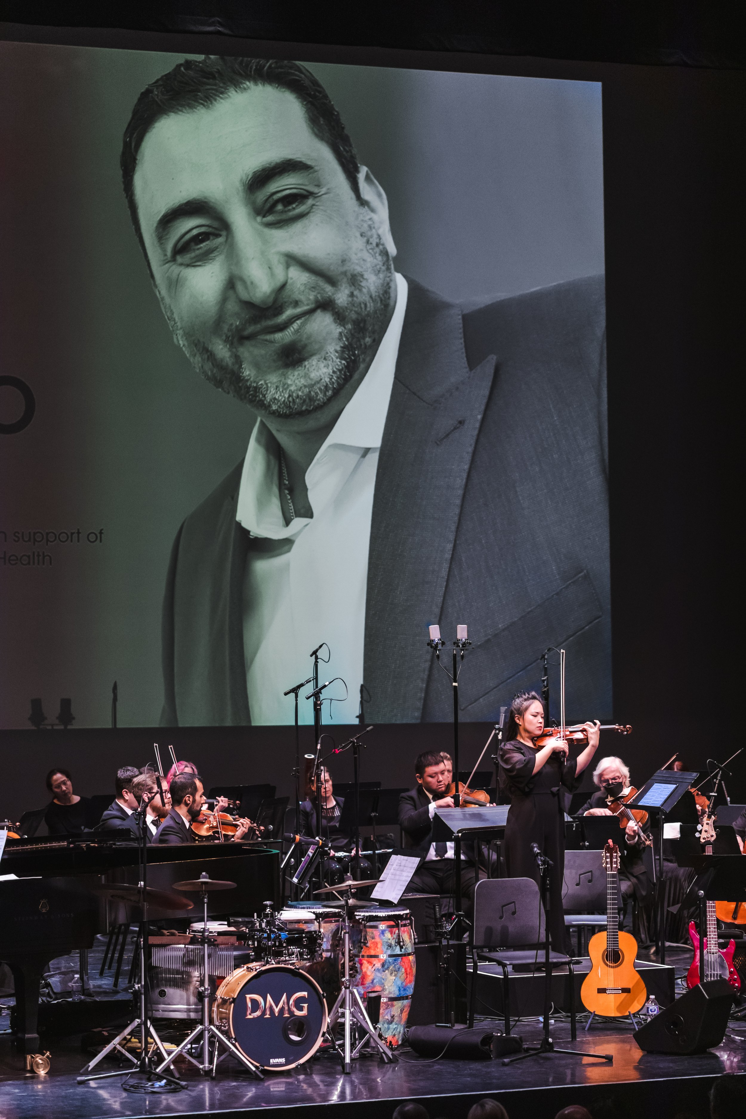 Orchestra performing on stage with a large screen showing a black-and-white photo of a man in a suit.