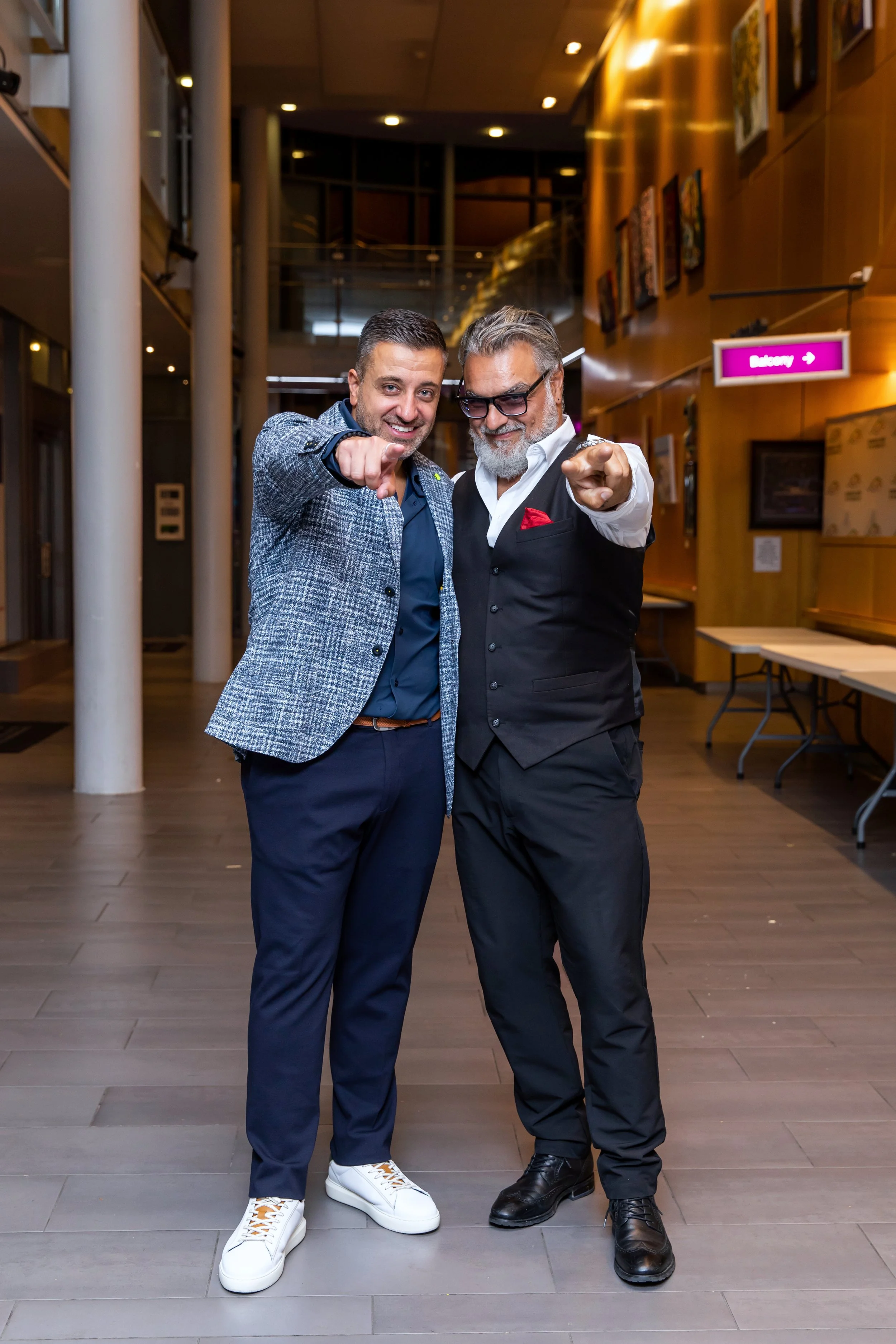 Two men are standing together indoors, pointing at the camera and smiling. One is wearing a gray checkered blazer with navy blue pants, white sneakers, and a blue shirt. The other man is dressed in black pants, a white shirt, a black vest, and black 