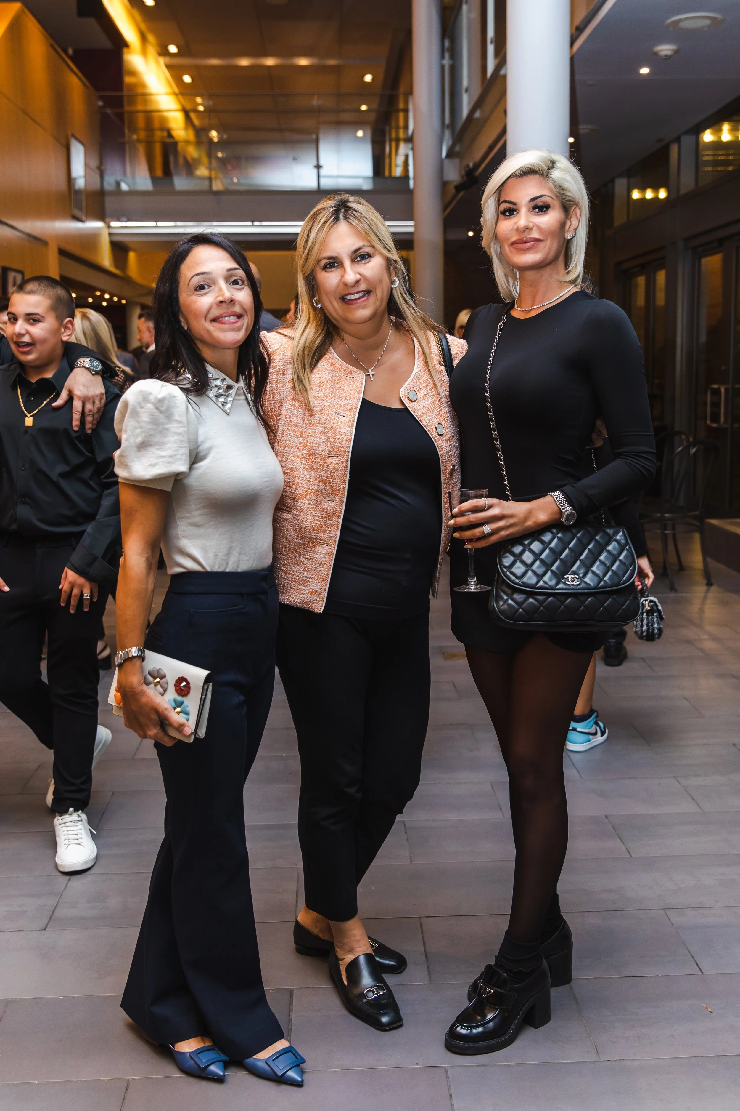 Three women posing at an indoor social event. They are dressed stylishly, and the woman on the right is holding a glass of wine and a quilted black purse. People can be seen in the background.