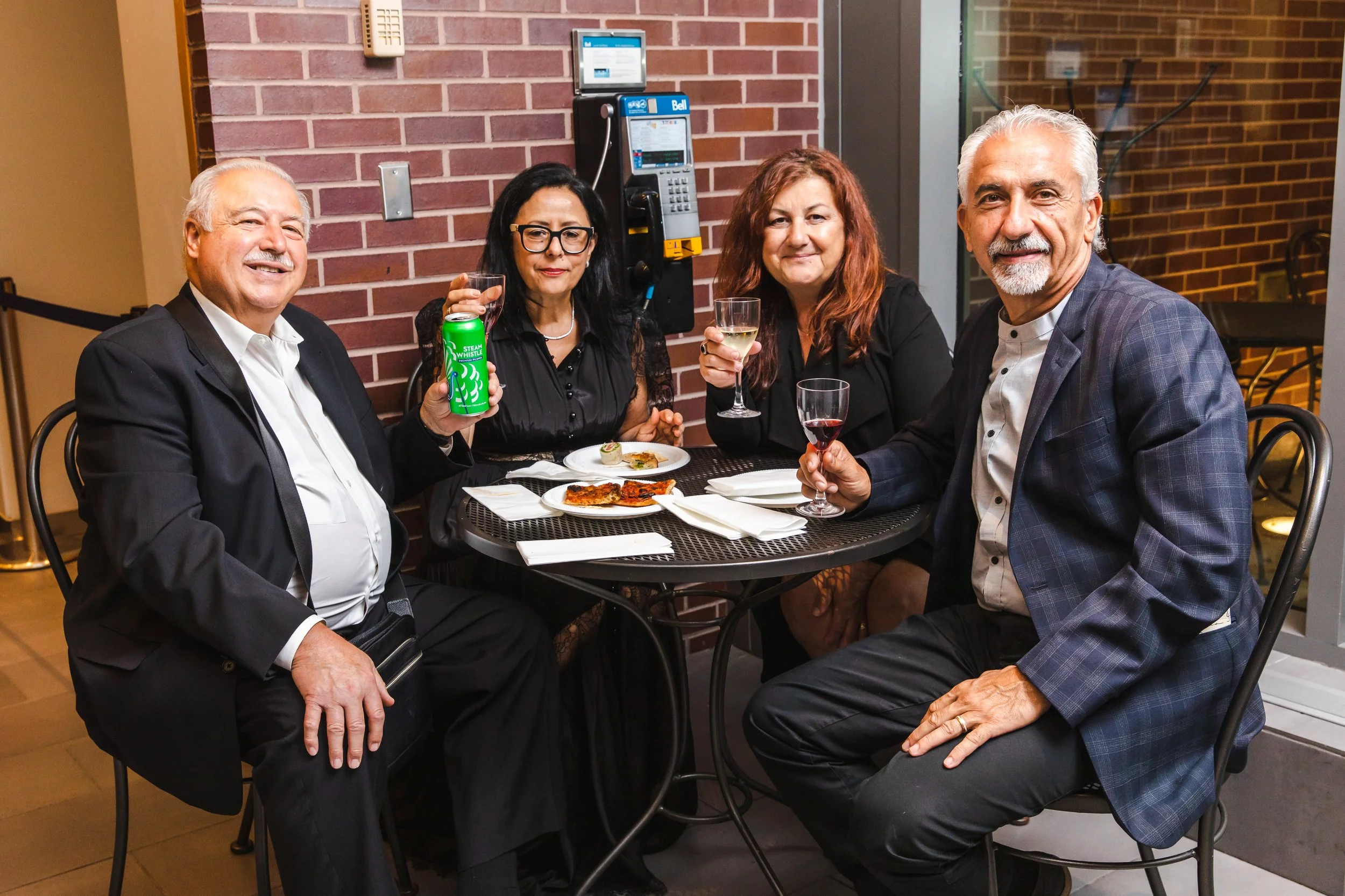 Group of four middle-aged adults, two men and two women, sitting at a restaurant table with wine and food, smiling at the camera, during evening or night.