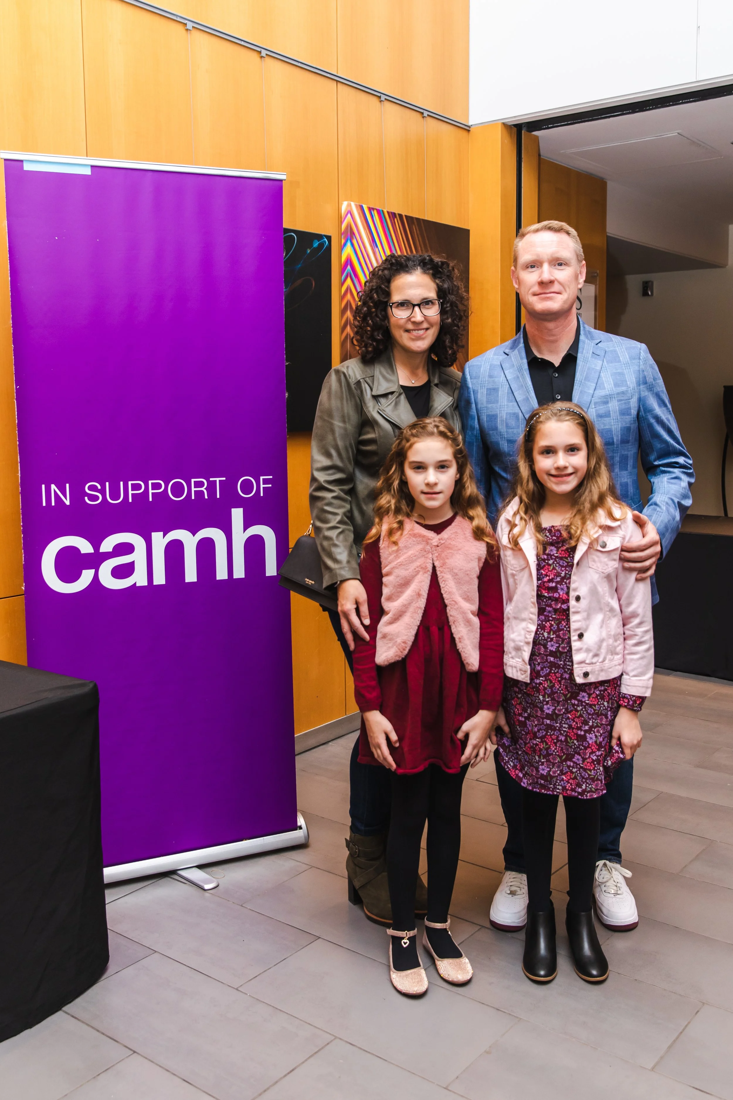 group of four people, two adults and two young girls, standing indoors near a purple banner that reads "IN SUPPORT OF camh". They are smiling and posing for a photo.
