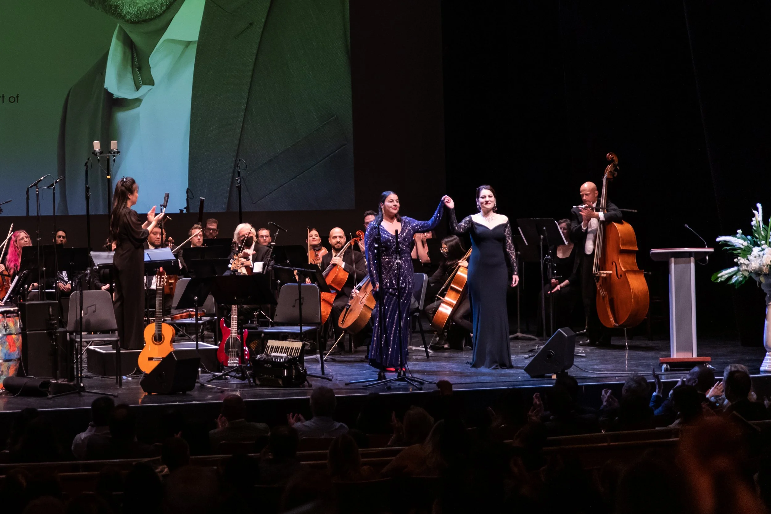 An orchestra performing on stage, with two women in elegant dresses sharing a bow at the center, surrounded by musicians playing string instruments, with a conductor and audience watching.
