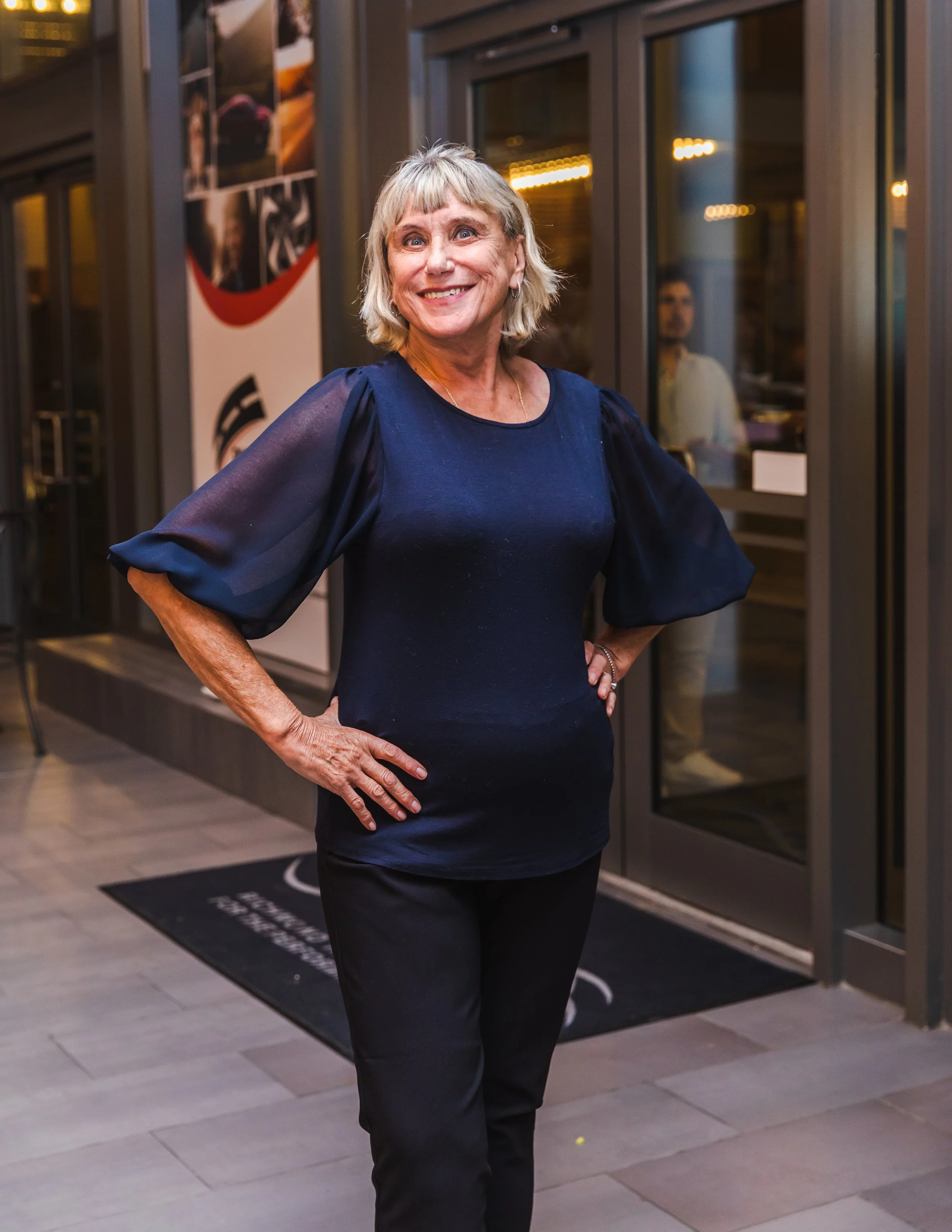 A woman with short, gray hair, wearing a navy blouse with puffed sleeves, standing outside a building, smiling with her hands on her hips.