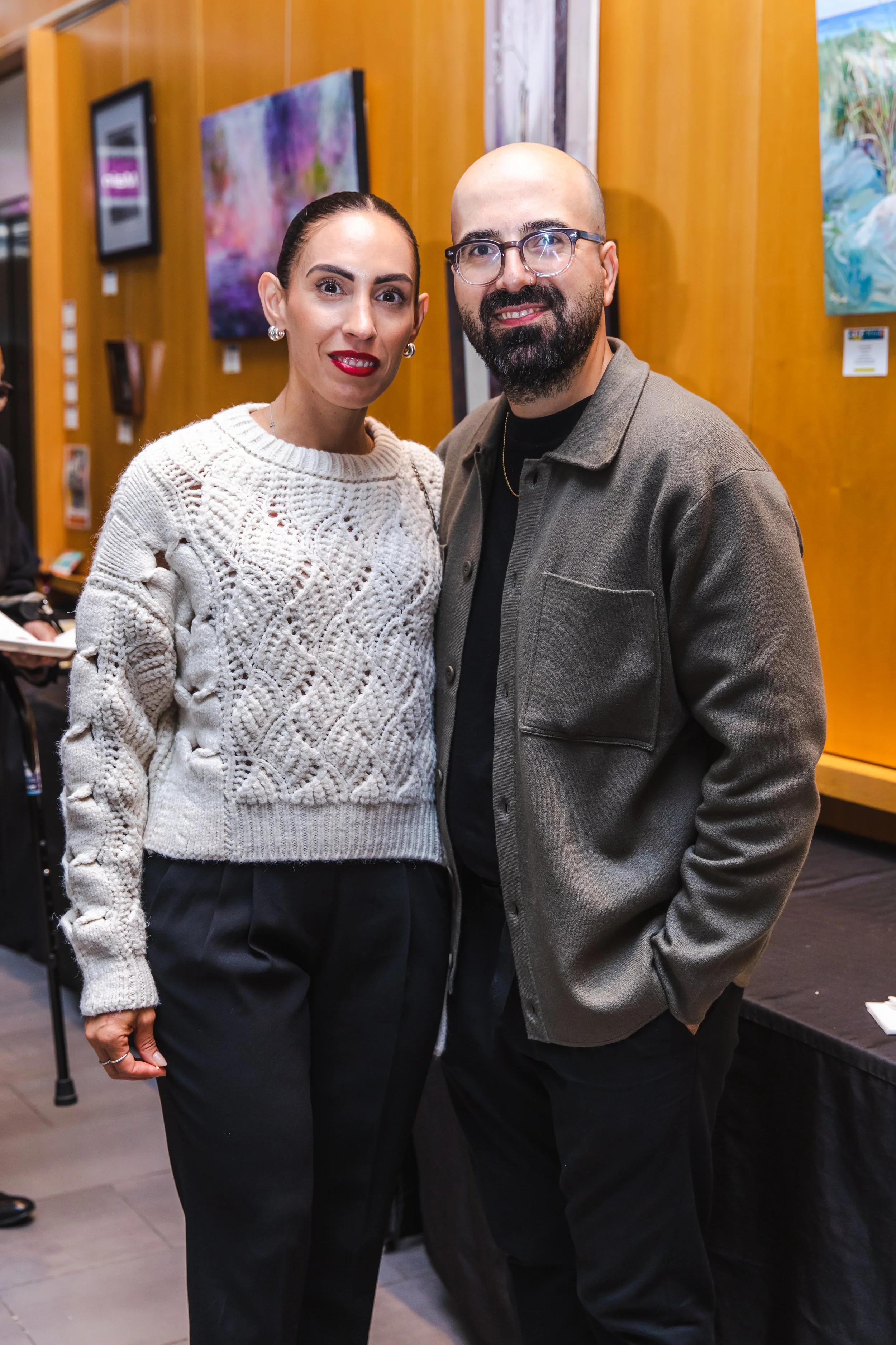 A woman in a white knit sweater and a man in glasses and a gray jacket standing together at an art gallery.