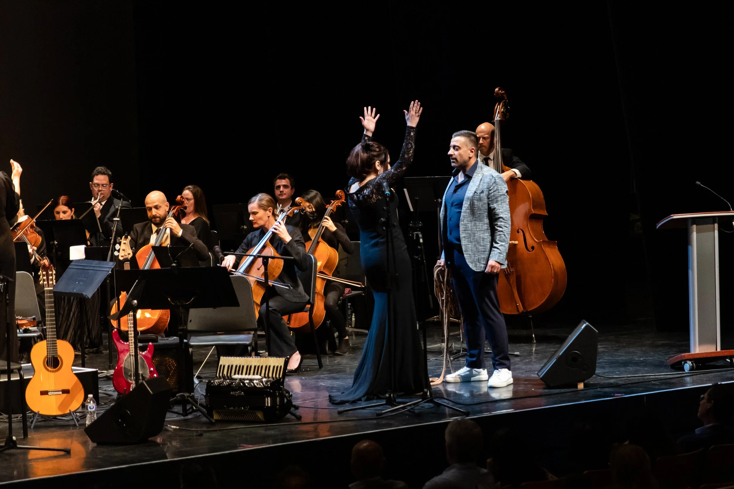 Orchestra performing on stage with conductor, musicians playing string instruments, and two individuals in conversation or interview at center stage.