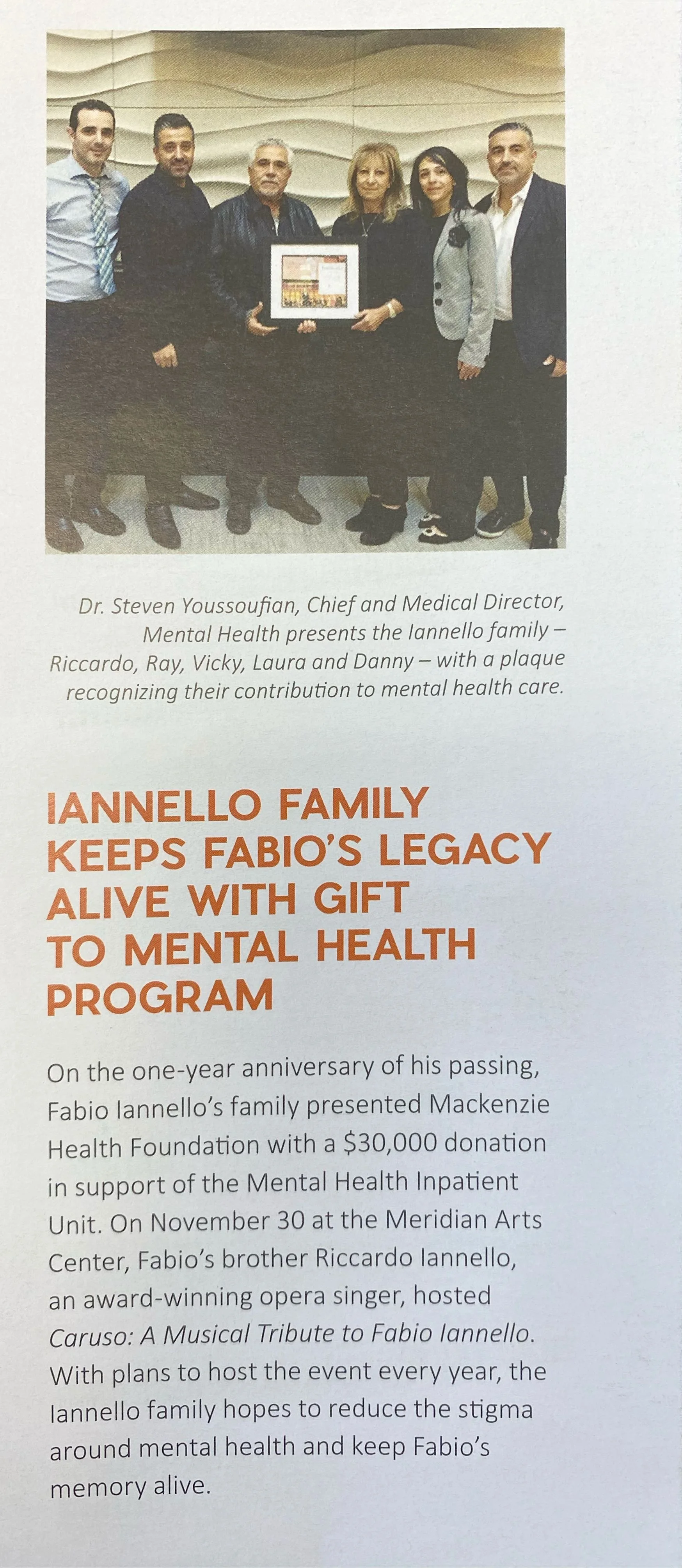 Group of six people standing together, holding a framed plaque, in front of a wavy textured wall.