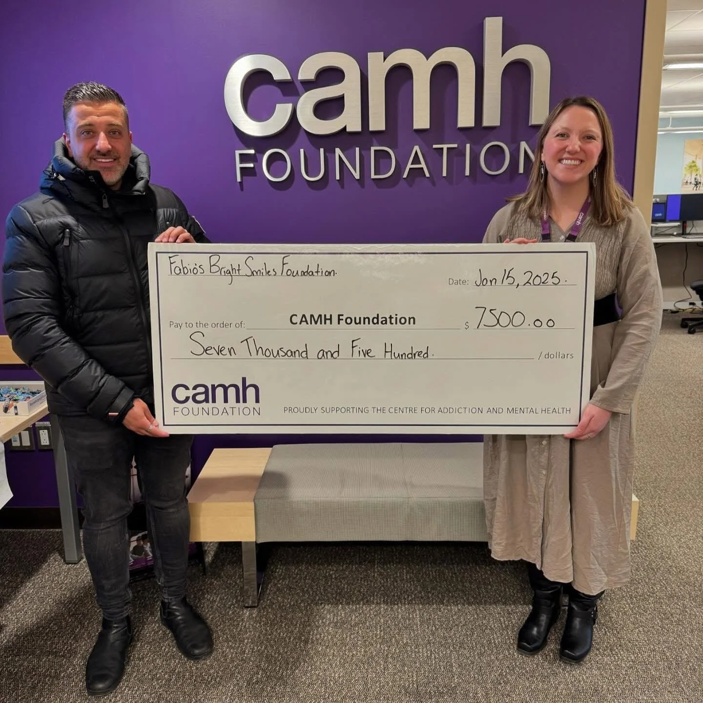Two people holding a large check in front of a purple wall with the words 'camh FOUNDATION'. The check is made out to CAMH Foundation for seven thousand five hundred dollars, dated January 15, 2025. The person on the left is wearing a black puffer jacket, and the person on the right is wearing a beige coat.