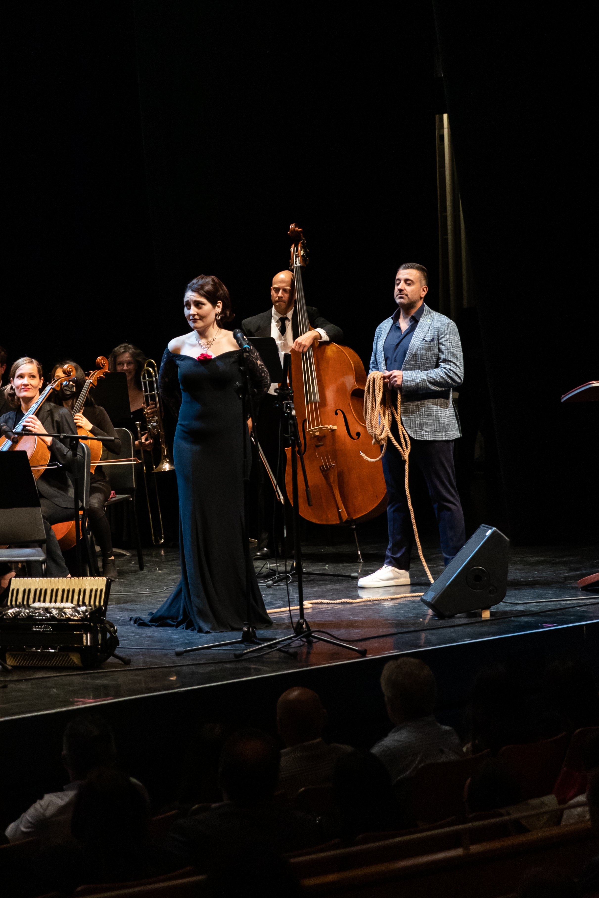 A woman in a long black dress speaking into a microphone on stage, with musicians and musical instruments behind her including a double bass, and a man holding a rope standing next to her at a live concert.