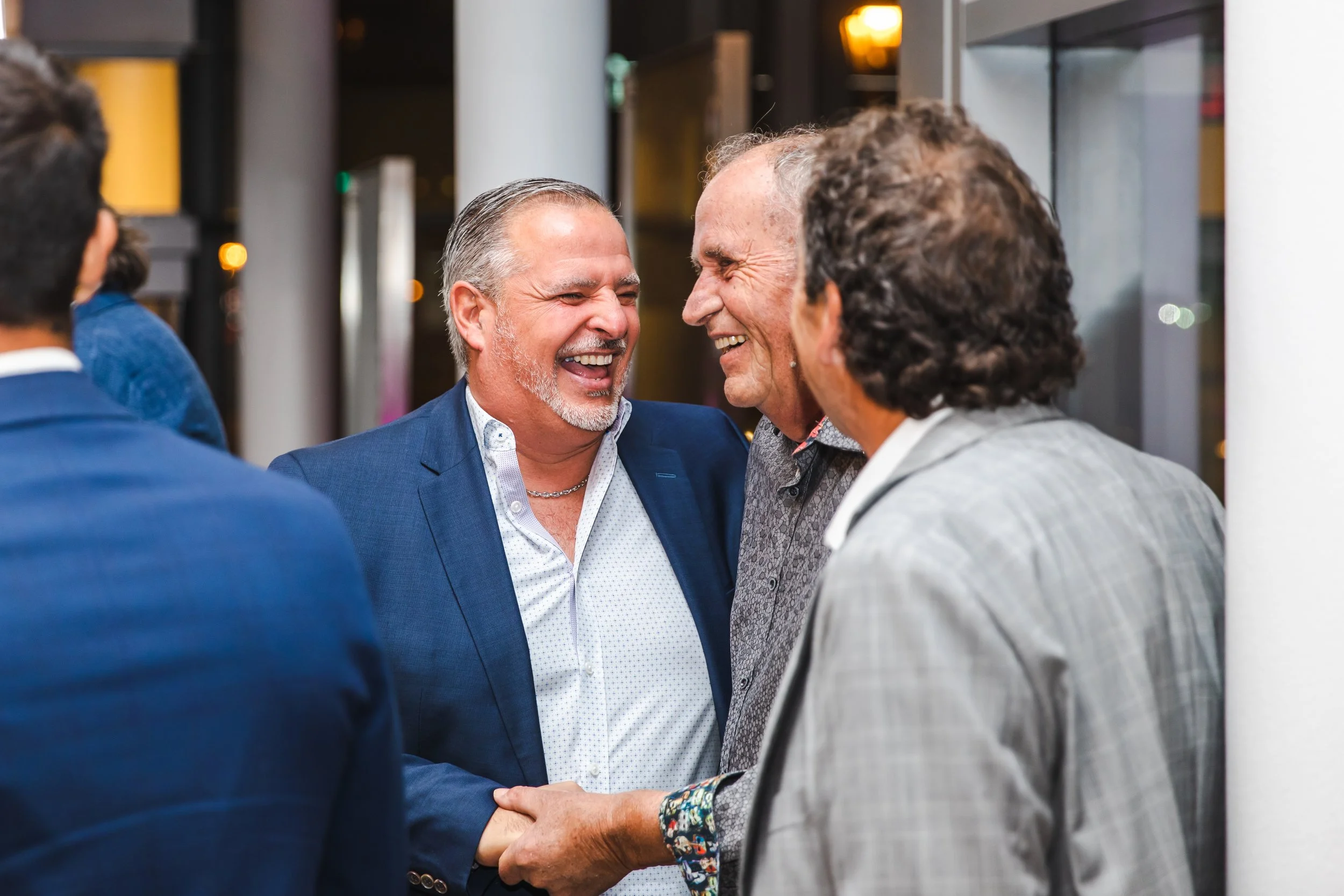 Four men engaged in conversation at a social event, smiling and laughing, with two of them shaking hands.